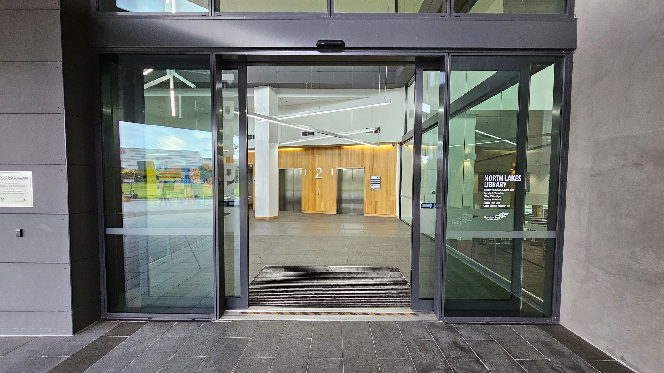 Automatic glass doors leading into a modern building. On the right door, "North Lakes Library" is written with opening hours. Inside, a wooden panelled wall with two lifts marked "1" and "2" is visible. The floor is tiled in dark grey, and a mat is placed at the entrance. The ceiling features geometric light fixtures. The exterior wall on the left is grey with a small sign.