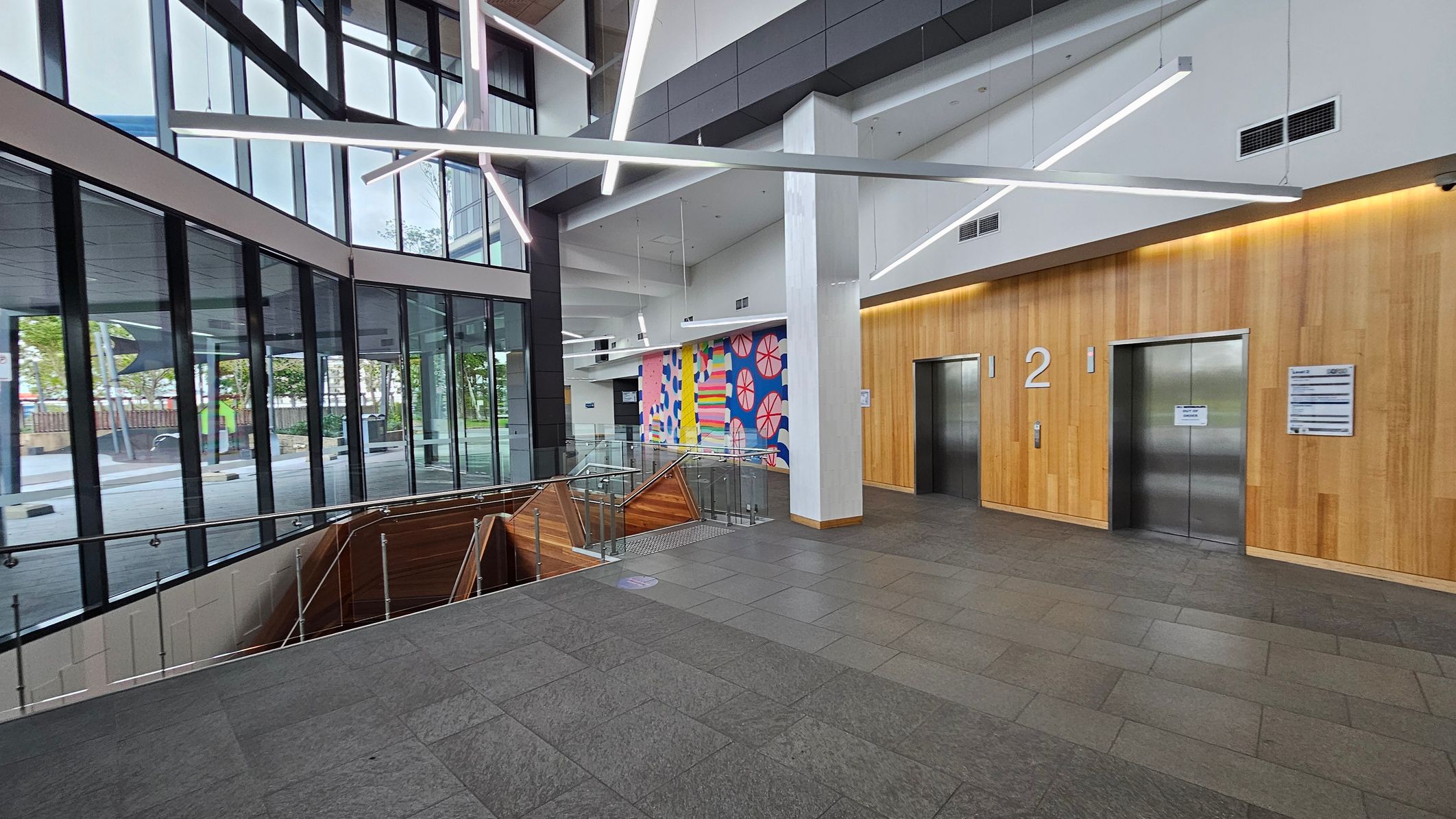 A modern interior with large glass windows on the left, allowing natural light to illuminate the space. In the centre, a staircase with wooden panelling descends, surrounded by glass railings. The right side features two lifts with a wooden wall backdrop, marked with the number "2". Above, geometric light fixtures hang from the ceiling. A colourful mural with abstract patterns is visible on the far wall. The floor is tiled in grey.