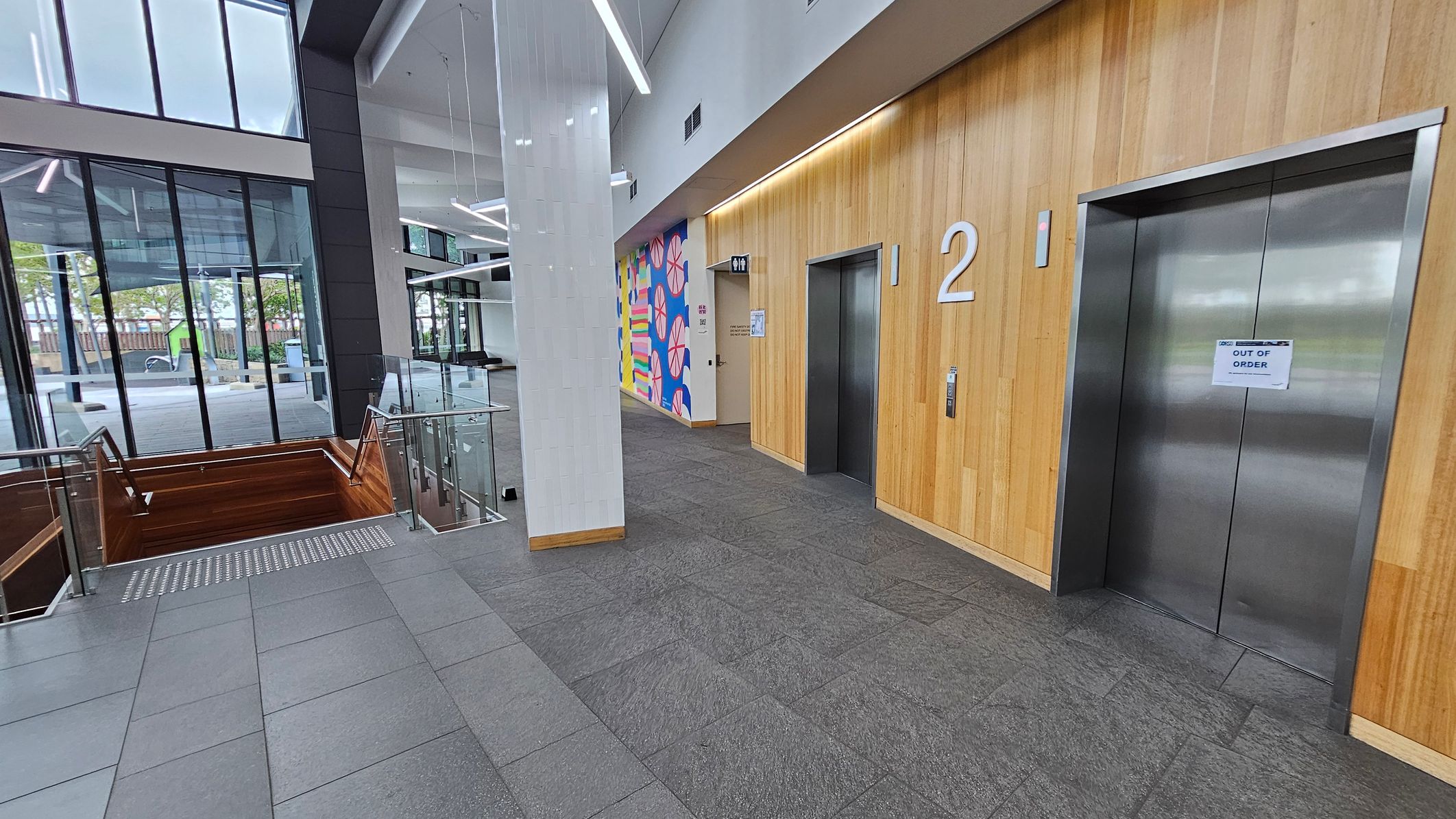 On the left, there are large glass windows with a view of the outside, showing trees and a pathway. In the middle, a staircase with wooden railings leads downwards, bordered by glass panels. On the right, two lift doors are set in a wooden panelled wall, with the number "2" above one. A sign on one lift reads "OUT OF ORDER." The floor is covered with grey tiles, and the ceiling features modern, angular light fixtures. A colourful mural is visible near the lifts.