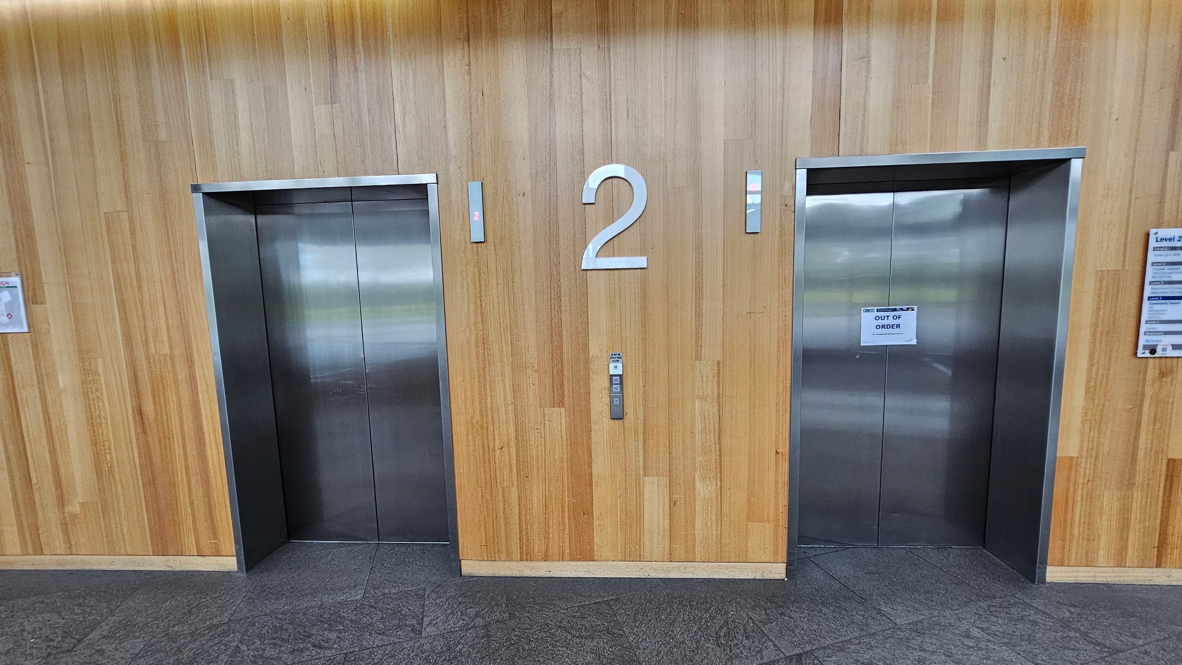Two lift doors set in a wooden panelled wall. The left lift is closed, while the right lift has a sign reading "OUT OF ORDER". A large number "2" is centred between the lifts, with smaller numbers "1" above each lift. The floor is covered with dark grey tiles. On the right, there is a directory sign mounted on the wall.