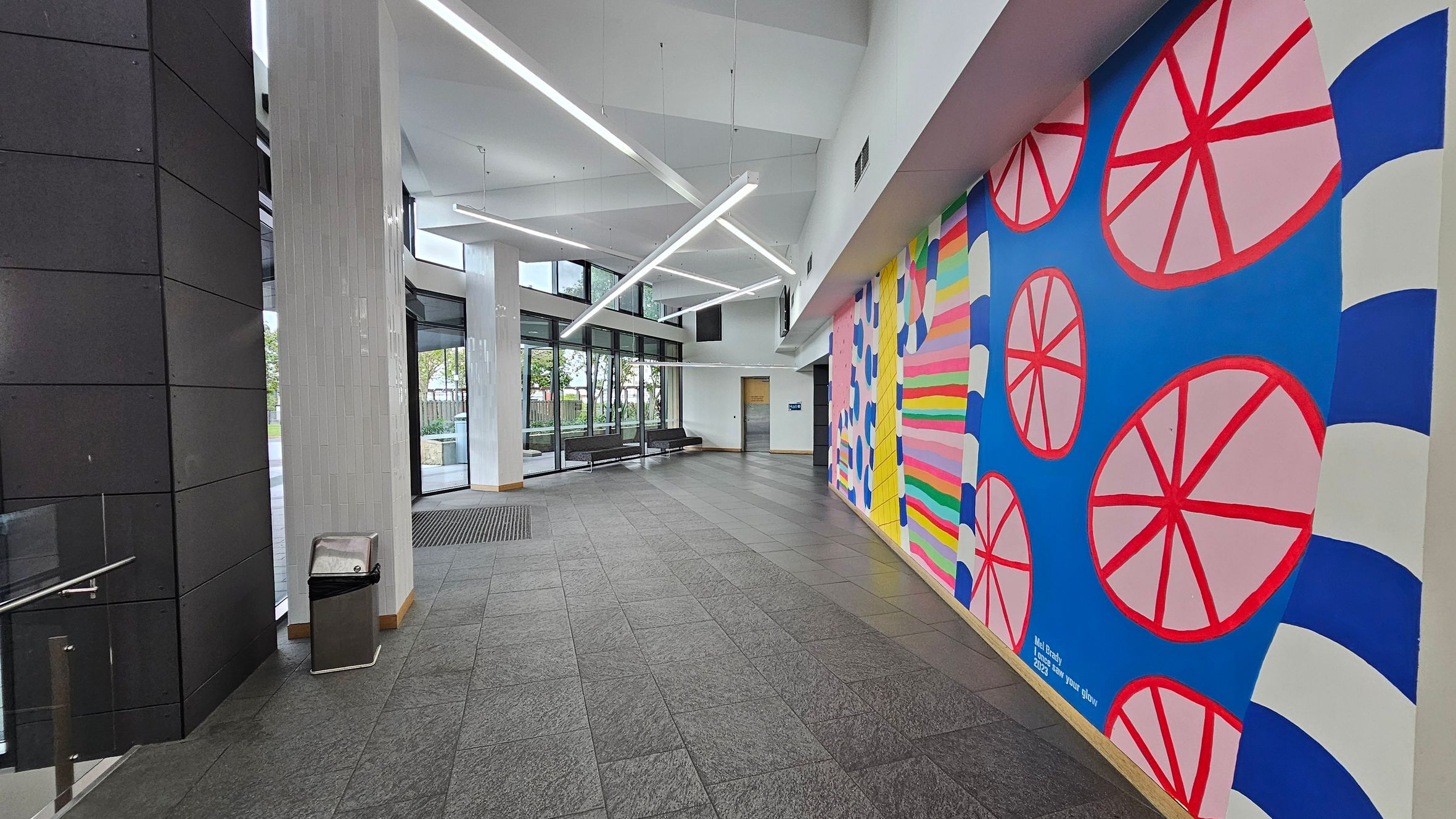 A modern interior space with a grey tiled floor and a high white ceiling featuring angular light fixtures. On the right, a vibrant mural with geometric patterns in blue, pink, and red. The left side has dark panelled walls and a glass entrance with large windows, allowing natural light to enter. A metal bin is positioned near the entrance, and there are black benches along the windowed wall.