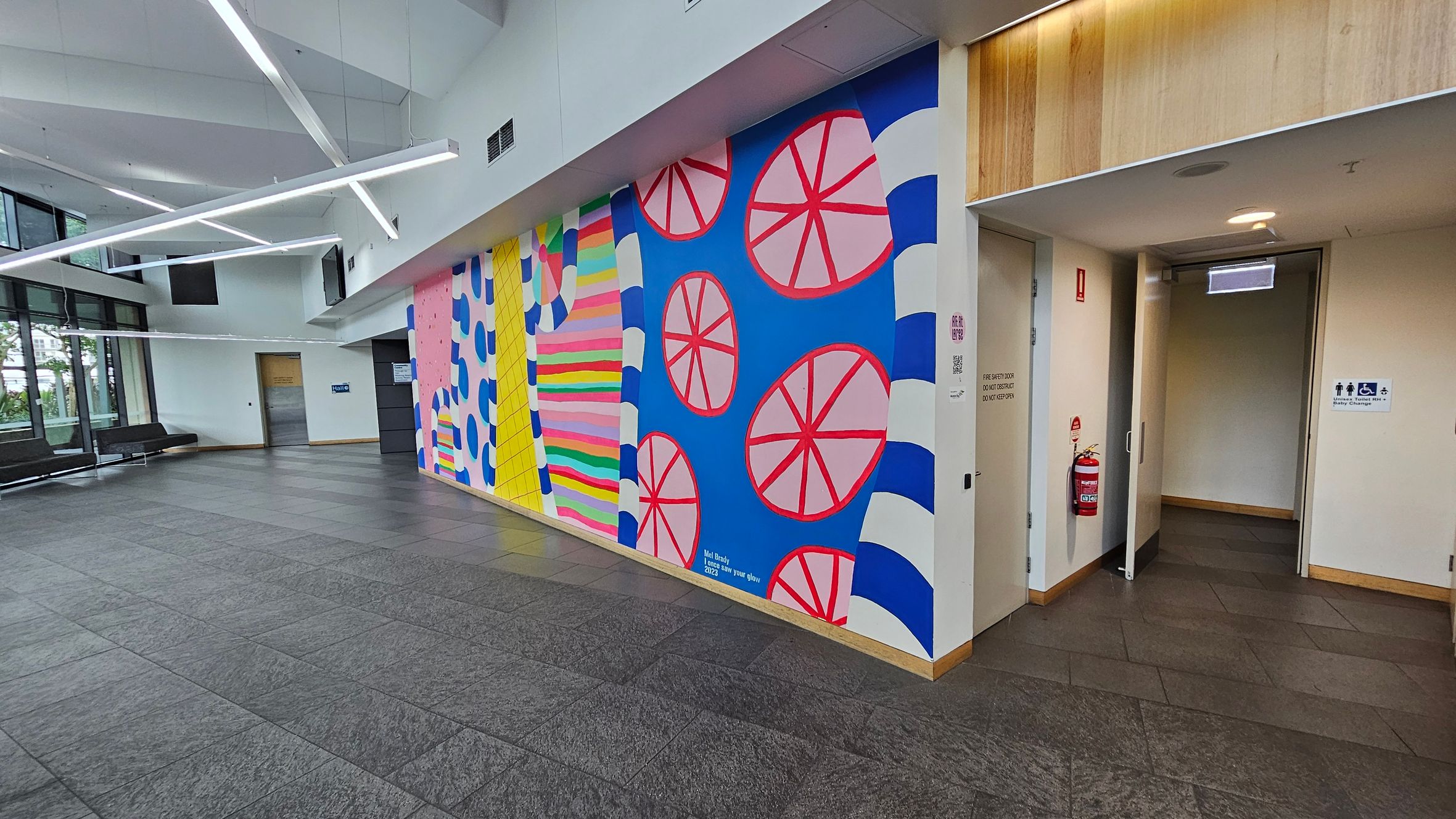 A spacious indoor area with a colourful mural on the right wall, featuring abstract patterns in pink, blue, yellow, and green. The mural includes circular shapes and stripes. To the right, there's a doorway leading to restrooms, marked with accessible signage. The floor is tiled in grey, and the ceiling has modern, angular light fixtures. On the left, large windows allow natural light in, with a row of black benches along the wall.