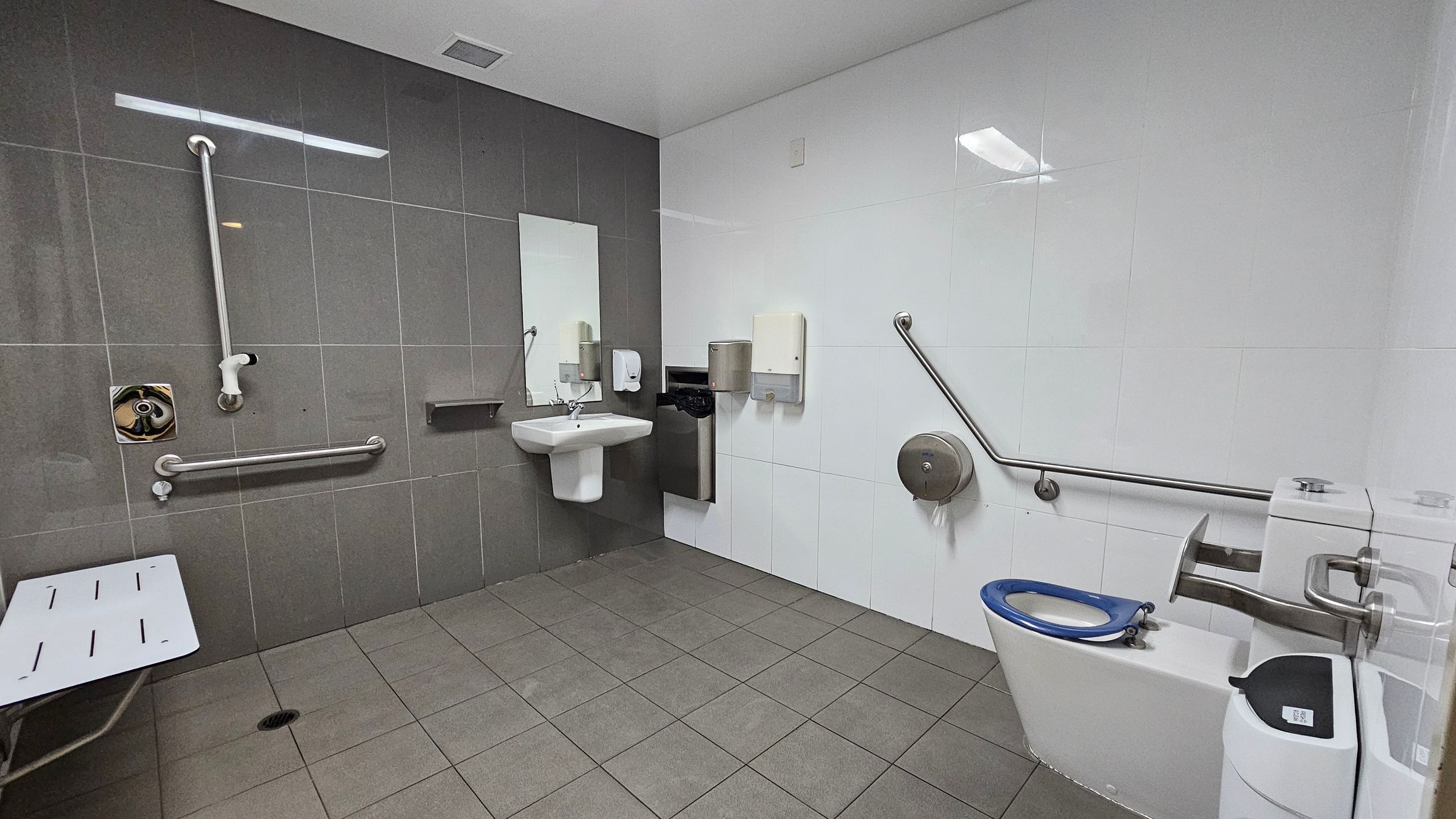 A public accessible bathroom with grey floor tiles and a white ceiling. On the left, there is a foldable shower seat and vertical and horizontal grab bars on the grey-tiled wall. In the centre, a white sink with a mirror above it is mounted on the wall, accompanied by a soap dispenser and a hand dryer. On the right, a toilet with a blue seat is equipped with horizontal and vertical grab bars, a large toilet paper dispenser, and a sanitary bin.