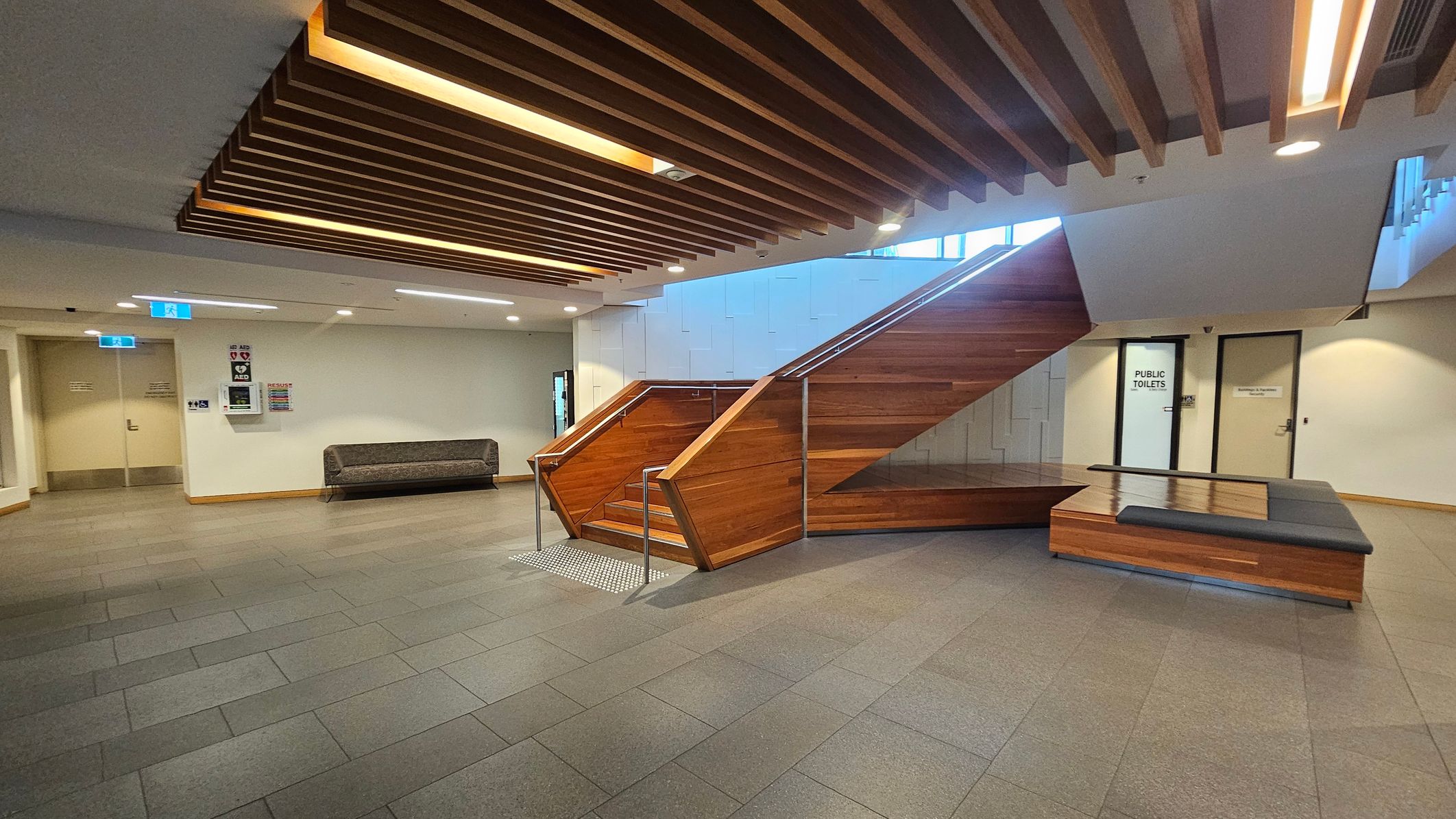 A modern interior space with a wooden staircase in the centre, featuring metal railings. To the left, there's a grey cushioned bench against a white wall with emergency signs and a door. On the right, a door marked "Public Toilets" is visible. The ceiling has wooden slats with integrated lighting, and the floor is tiled in grey. A wooden bench with a cushioned top is positioned near the staircase.