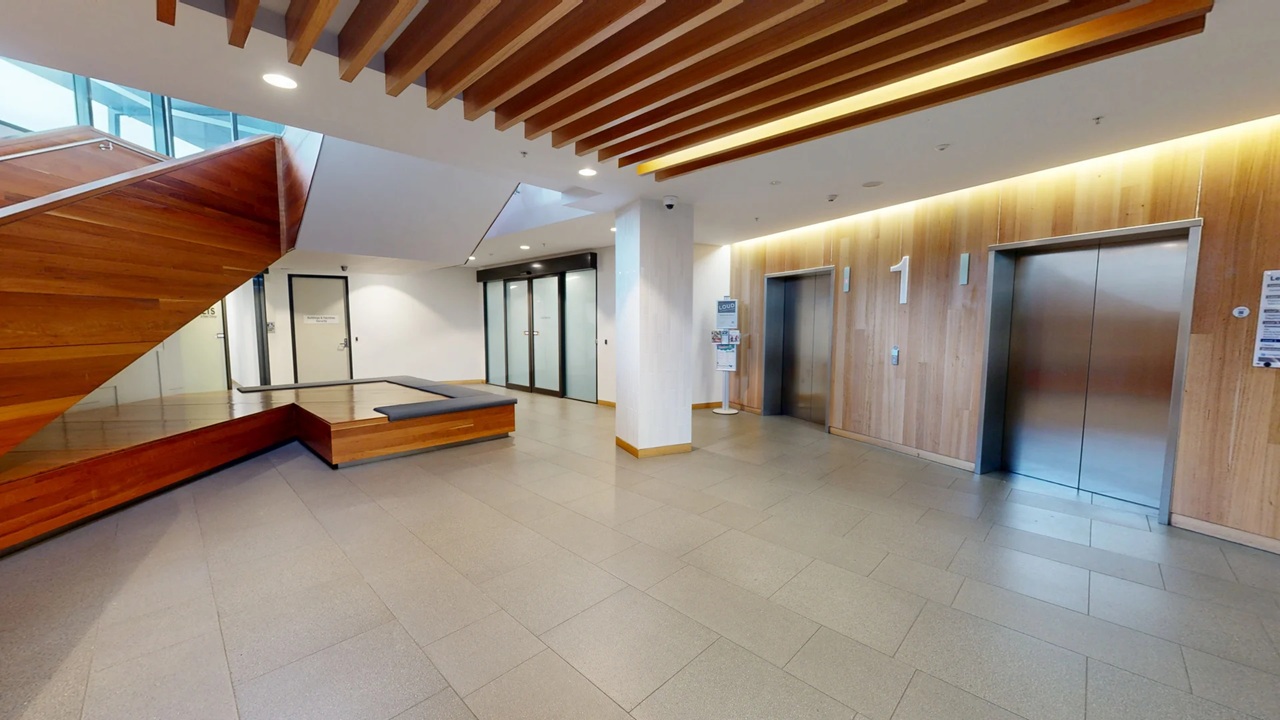 A modern interior space with a wooden staircase on the left, featuring a glass railing. The floor is tiled in a light colour. In the centre, there is a seating area with a wooden bench. On the right, two lift doors are set into a wooden panelled wall, with the number "1" visible above them. The ceiling has wooden slats and recessed lighting. A water dispenser is positioned near the lifts.
