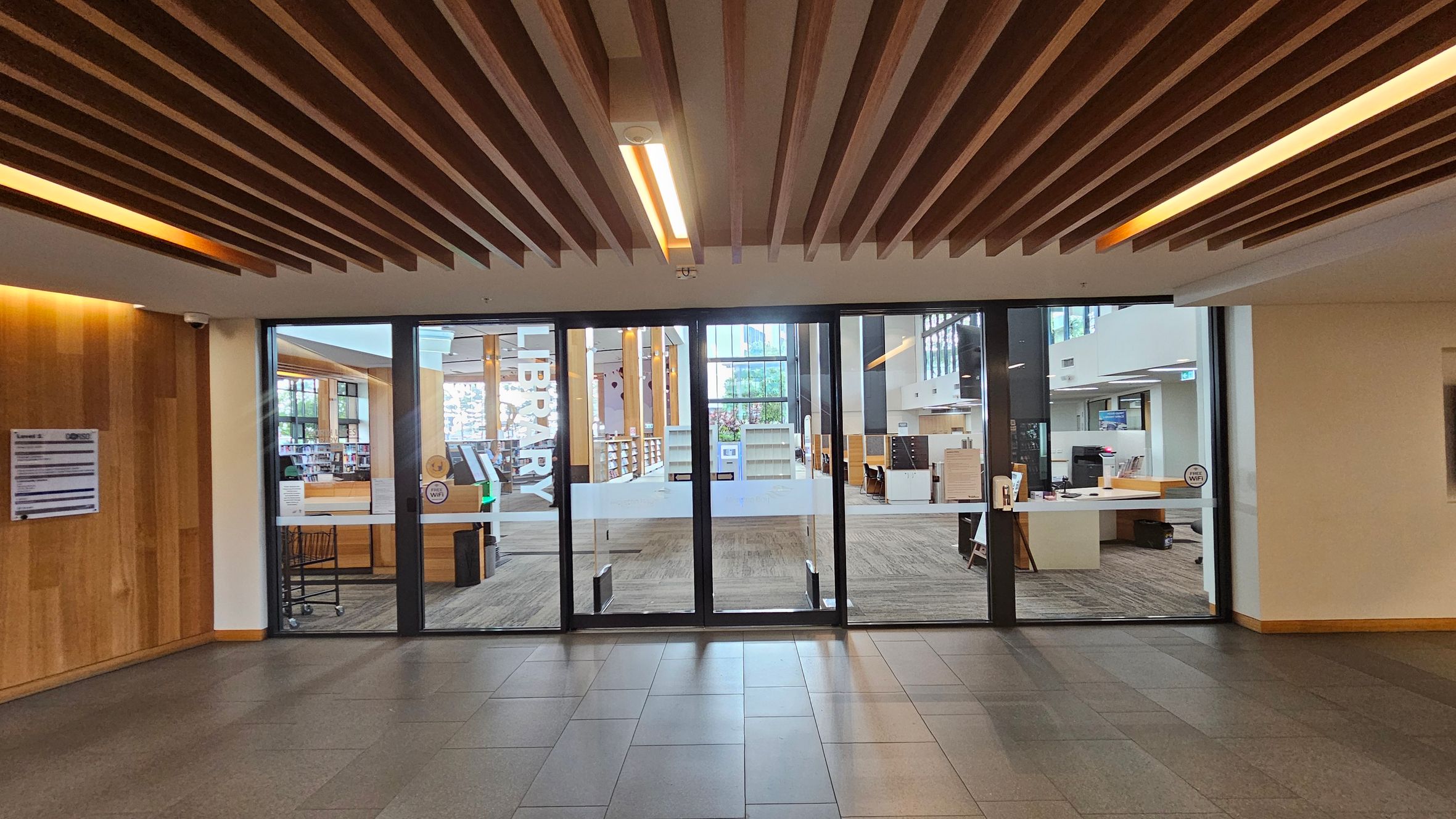 A modern library entrance with large glass doors in the centre, displaying the word "LIBRARY" vertically. Inside, there are bookshelves and a reception desk visible. The floor is tiled in a dark grey, and the ceiling features wooden slats with integrated lighting. On the left, a wooden wall with a notice board is present. The interior beyond the doors has a carpeted floor and large windows allowing natural light.