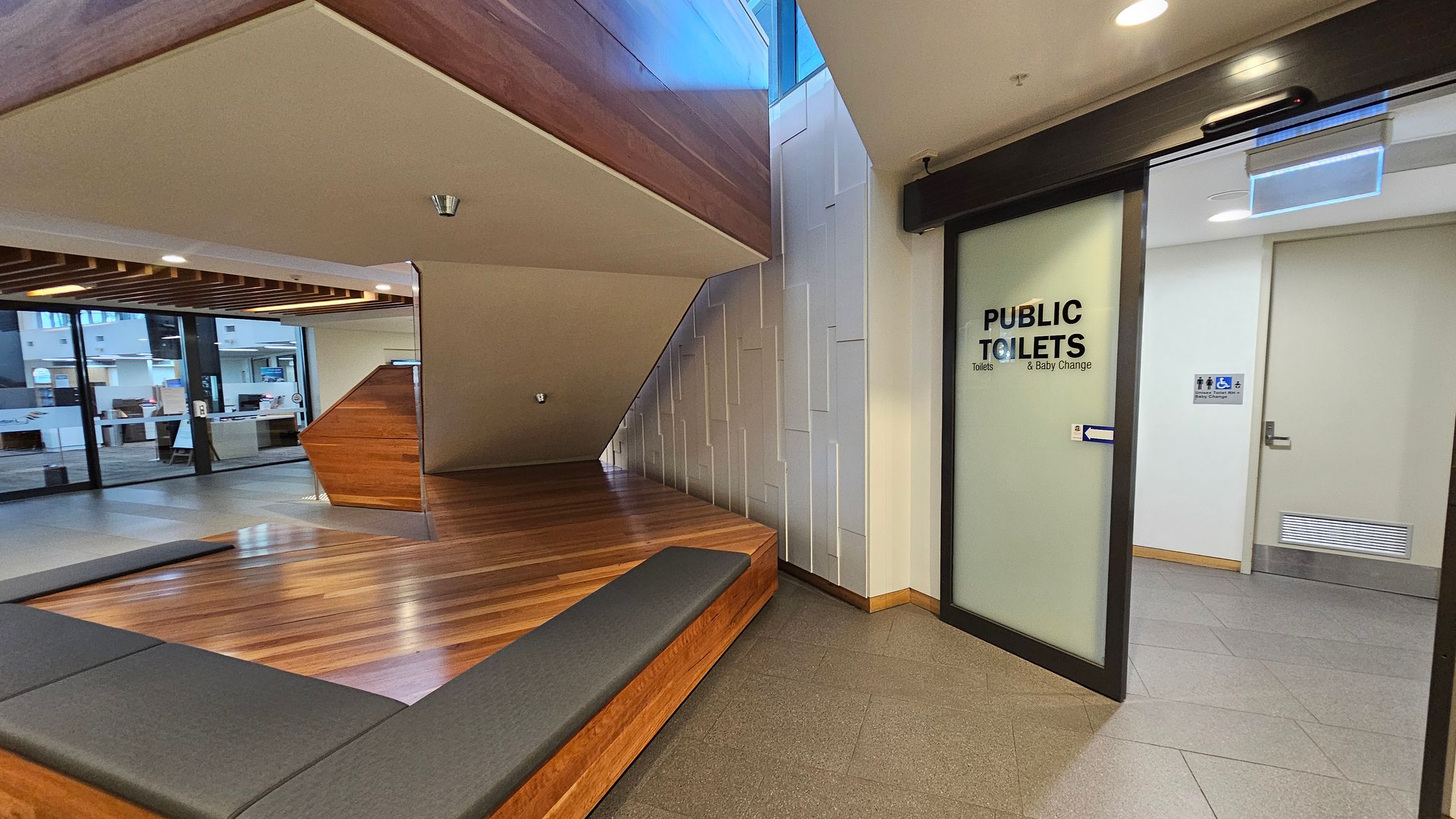 A modern interior space with a wooden bench and flooring on the left, leading to a seating area. The ceiling has recessed lighting and wooden beams. On the right, there is a frosted glass door with "PUBLIC TOILETS" written on it, leading to a restroom. The wall beside the door has a textured design. In the background, large windows reveal an exterior view. The floor is a mix of wood and grey tiles.