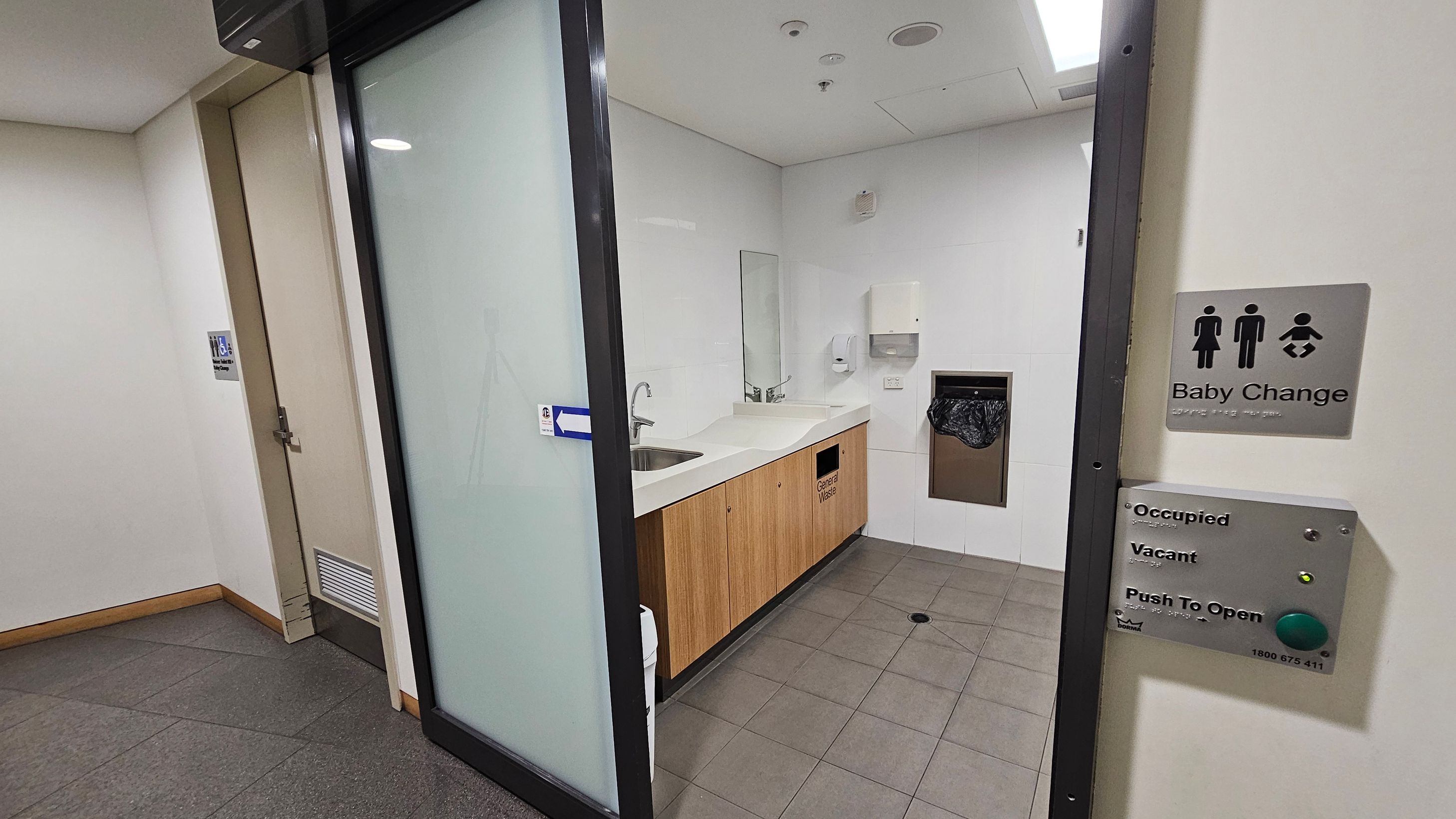 A baby changing room with a wooden cabinet and a white countertop on the left, featuring a sink and a mirror above. A waste bin is integrated into the cabinet. On the right, a sign indicates "Baby Change" with symbols for male, female, and baby. Below, there are buttons for occupancy status and a "Push to Open" button. The floor is tiled, and the ceiling has recessed lighting. A closed door is visible in the background.