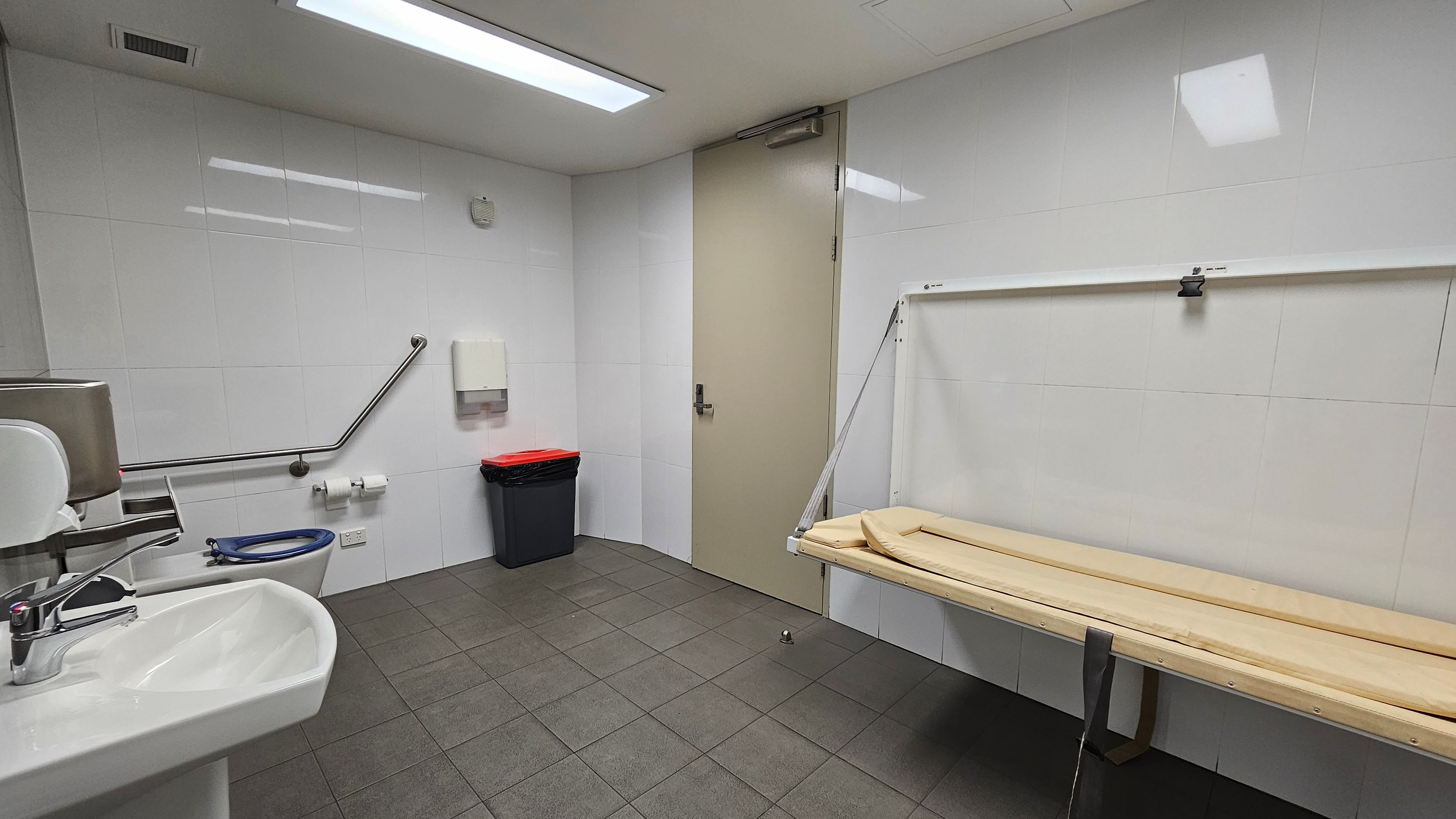 A clean, white-tiled bathroom with a grey tiled floor. On the left, there's a white sink with a chrome tap and a wall-mounted soap dispenser. A toilet with a blue seat is next to it, equipped with a grab rail. In the centre, a black bin with a red lid is beneath a paper towel dispenser. On the right, a beige door and a foldable changing table are visible. The ceiling has a rectangular fluorescent light.