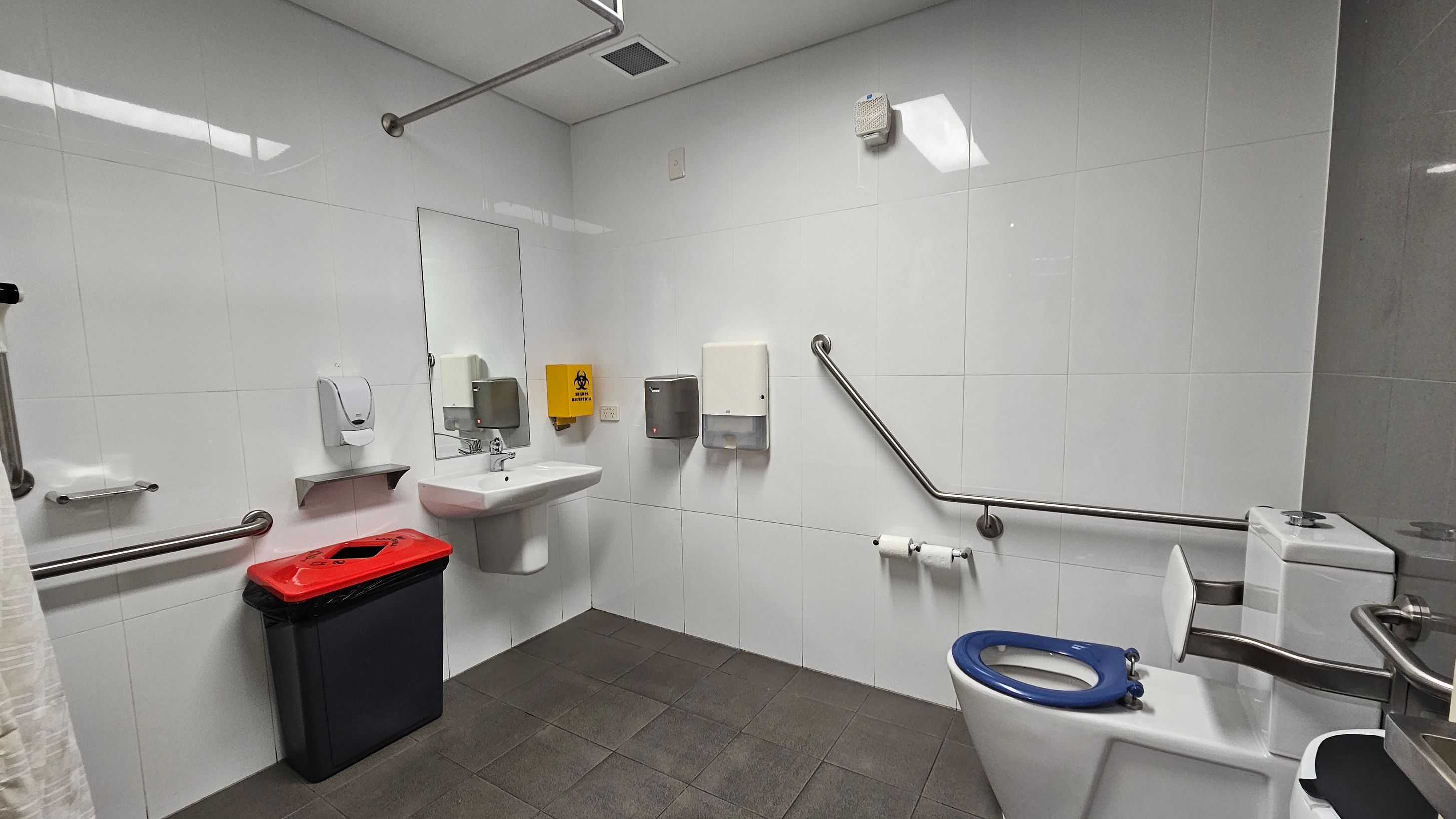 A public restroom with white tiled walls and a grey tiled floor. On the left, a red-lidded bin is beneath a soap dispenser and a hand dryer. A large mirror is above a white sink. In the centre, a yellow biohazard disposal unit and two paper towel dispensers are mounted on the wall. On the right, a toilet with a blue seat is equipped with support rails. A toilet roll holder is attached to the wall. The ceiling is white with a vent and a light fixture.