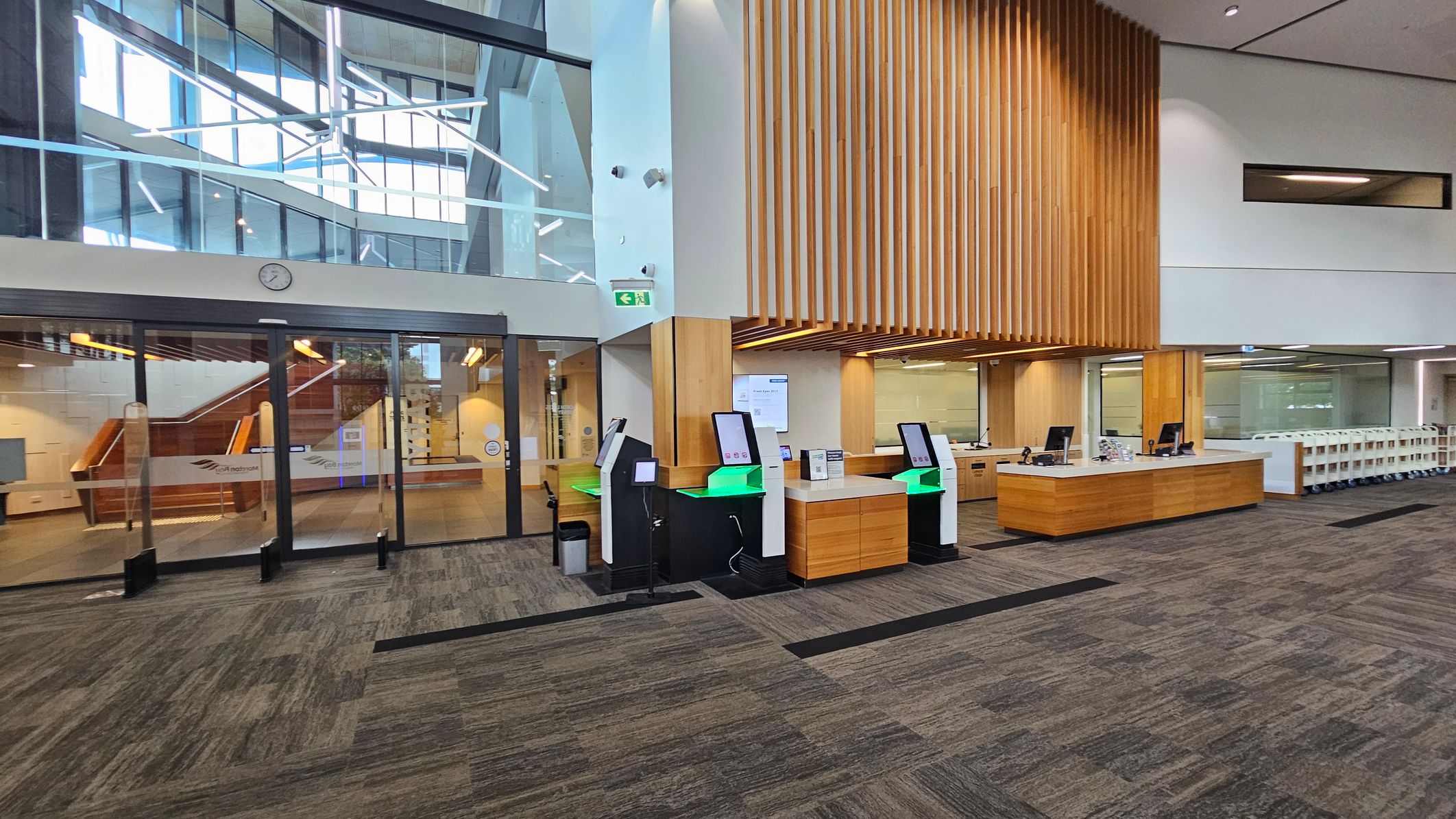 A modern interior with a spacious layout. On the left, there are large glass doors with a clock above, leading to a staircase with wooden railings. In the centre, several self-service kiosks with green lighting are positioned on a patterned carpeted floor. On the right, a long wooden counter with computer stations is set against a wall with vertical wooden slats. The ceiling is high with recessed lighting, and the area is well-lit with natural light from large windows.