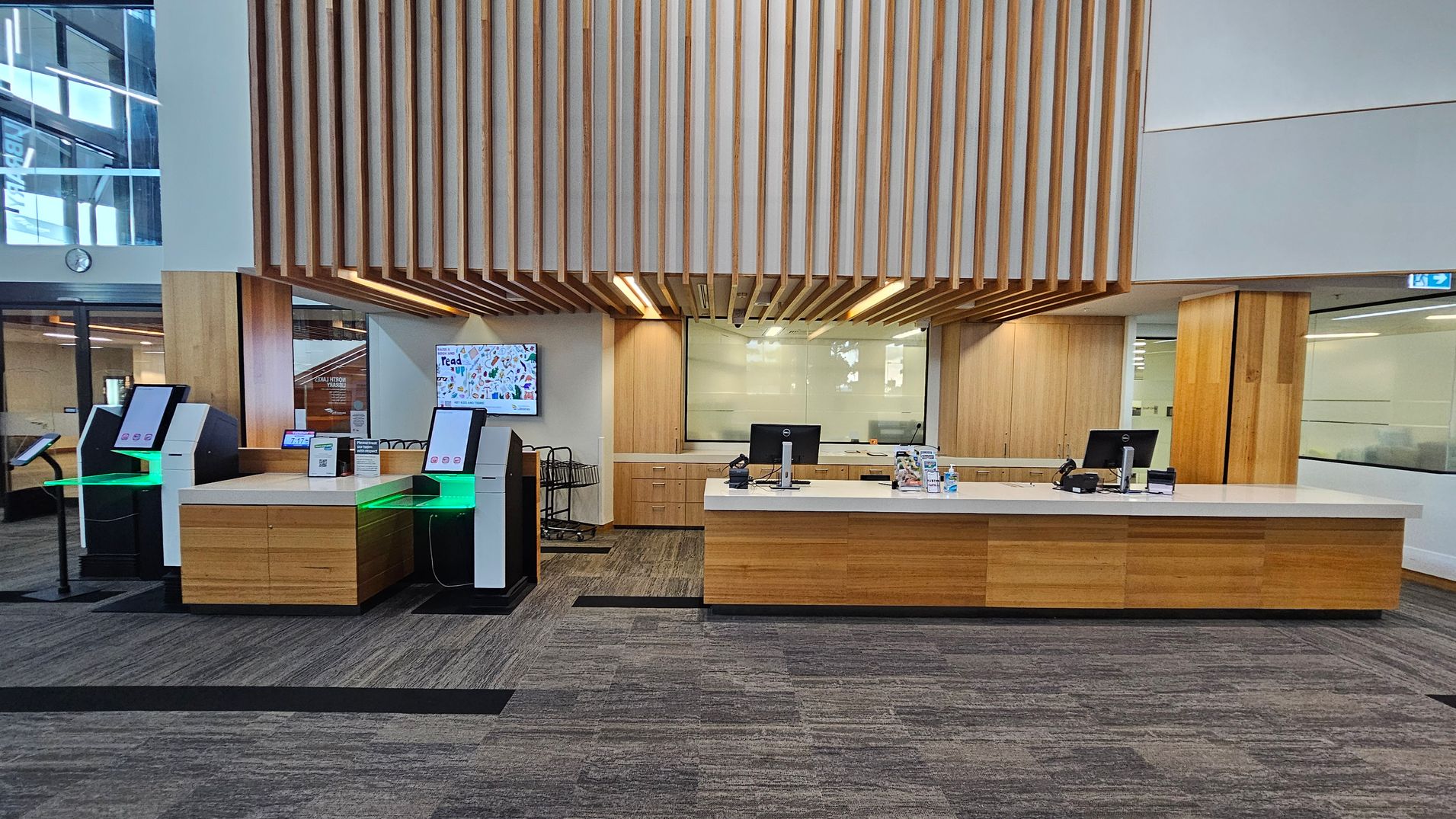 A modern interior with a reception area. On the left, there are two self-service kiosks with green lighting. In the centre, a wooden counter with a computer and various items on top. Behind the counter, a glass partition and wooden panelling on the walls. Above, a ceiling feature with wooden slats. The floor is carpeted in a grey pattern. On the left wall, a digital screen displays colourful graphics.