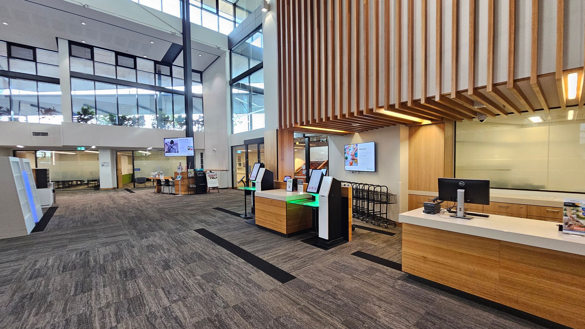 A modern interior with high ceilings and large windows. On the right, there is a wooden reception desk with a computer and brochures. Above, wooden slats form a decorative ceiling feature. In the centre, two self-service kiosks with green lighting are positioned on a wooden base. On the left, a glass-walled meeting room is visible, and a small seating area with brochures is nearby. The floor is carpeted in a grey and purple pattern.