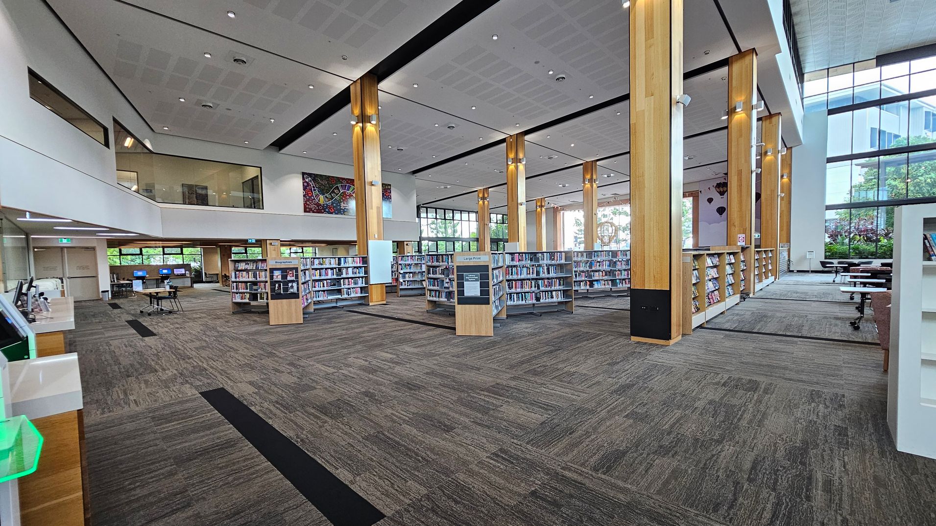 A spacious library interior with tall wooden columns and a high ceiling. In the centre, there are several bookshelves filled with books. To the left, there are computer stations and a service desk. Large windows on the right allow natural light to illuminate the space, with tables and chairs near the windows. The floor is carpeted in a grey pattern, and the ceiling features recessed lighting. A colourful artwork is visible on the far wall.