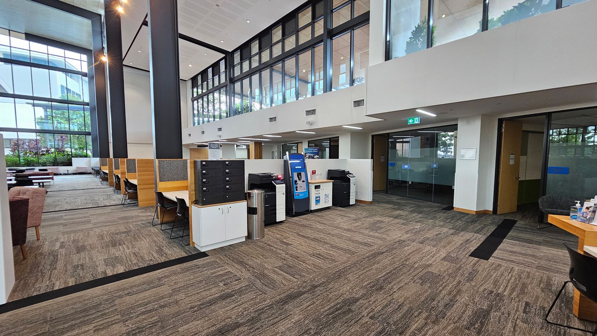 A spacious, modern interior with a high ceiling and large windows on the left, allowing natural light to flood in. On the right, there are glass-walled offices and a row of printers and a kiosk. In the middle, there are workstations with chairs and dividers. The floor is carpeted in a neutral tone, and the ceiling features recessed lighting. The background includes greenery visible through the windows, adding a touch of nature to the space.
