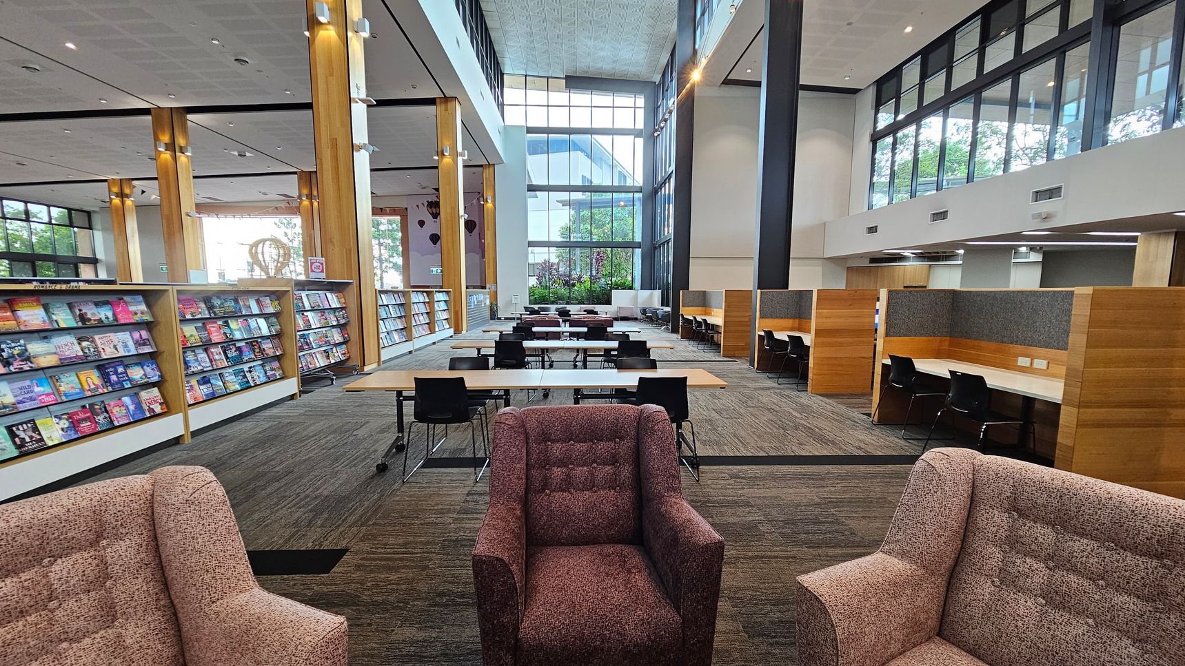 A spacious library interior with high ceilings and large windows. On the left, there are shelves filled with colourful books. In the centre, long tables with black chairs are arranged for study. On the right, there are individual study booths with wooden partitions. The foreground features two upholstered armchairs. The floor is carpeted in a neutral tone, and the ceiling has a modern design with recessed lighting.