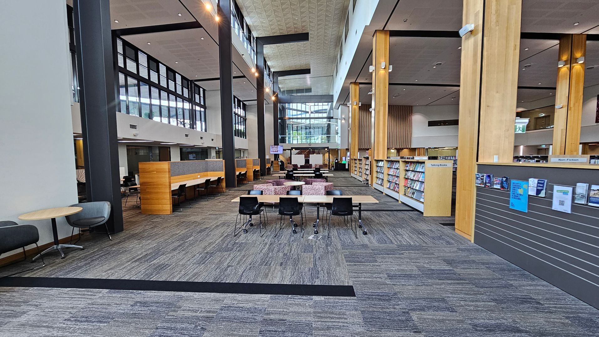 A spacious library interior with a high ceiling. On the left, there are small round tables with grey chairs and a long wooden partition with study desks. In the centre, there are tables with black chairs and a seating area with patterned sofas. On the right, bookshelves labelled "Non-Fiction" and "Talking Books" are visible, along with a wall displaying notices. The floor is carpeted in a mix of grey and purple tones, and large windows provide natural light.