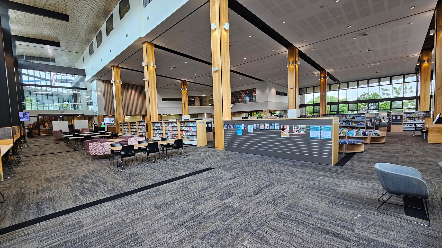 A spacious library interior with a high ceiling and large windows in the background. The floor is covered with grey and brown patterned carpet. On the left, there are study tables with chairs and a row of bookshelves. In the centre, wooden columns support the ceiling, and more bookshelves are visible. On the right, a grey chair is placed near a partition displaying notices. The area is well-lit with natural light streaming through the windows.