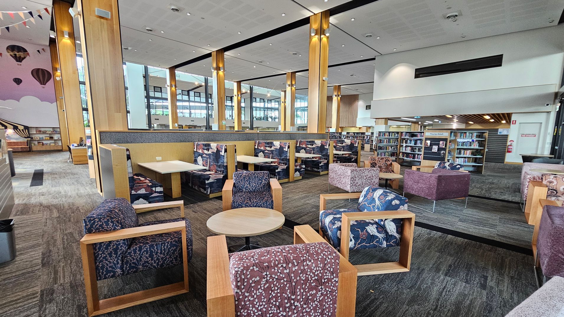 A spacious library interior with a modern design. In the foreground, there are several cushioned chairs with wooden frames and a round wooden table. The middle area features booth seating with patterned upholstery and small tables. The background has tall wooden columns and large windows allowing natural light. Bookshelves are visible on the right, with a sign indicating a "Young Adults" section. The ceiling is high with recessed lighting, and the floor is carpeted in a neutral tone.