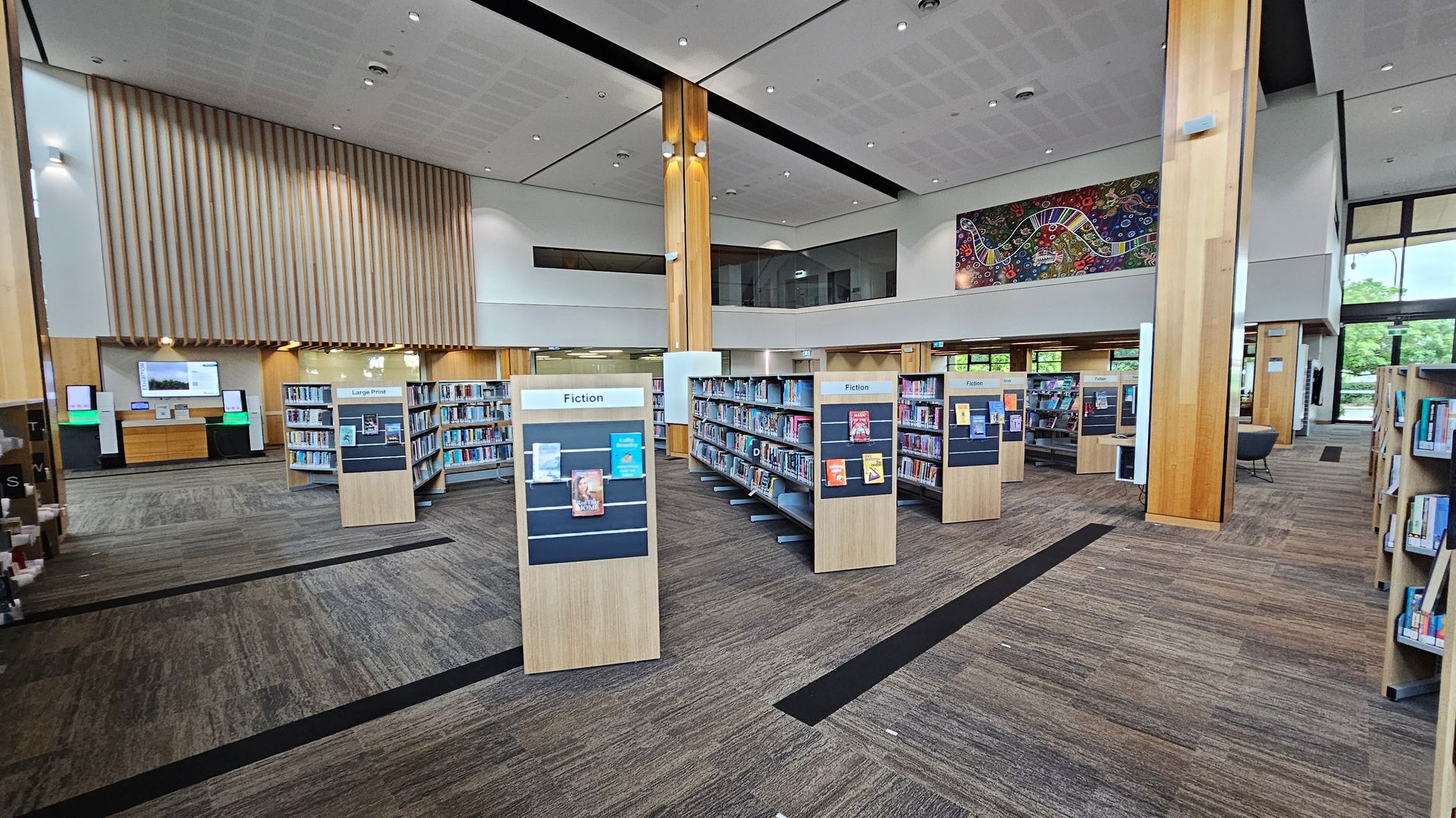 A spacious library interior with a high ceiling and wooden beams. In the foreground, there are several bookshelves labelled "Fiction" and "Large Print," filled with books. The floor is carpeted in a mix of grey and brown tones. In the background, a large, colourful mural adorns the wall above more bookshelves. To the left, there are digital kiosks and a wooden panelled wall. Large windows on the right allow natural light to enter, with greenery visible outside.