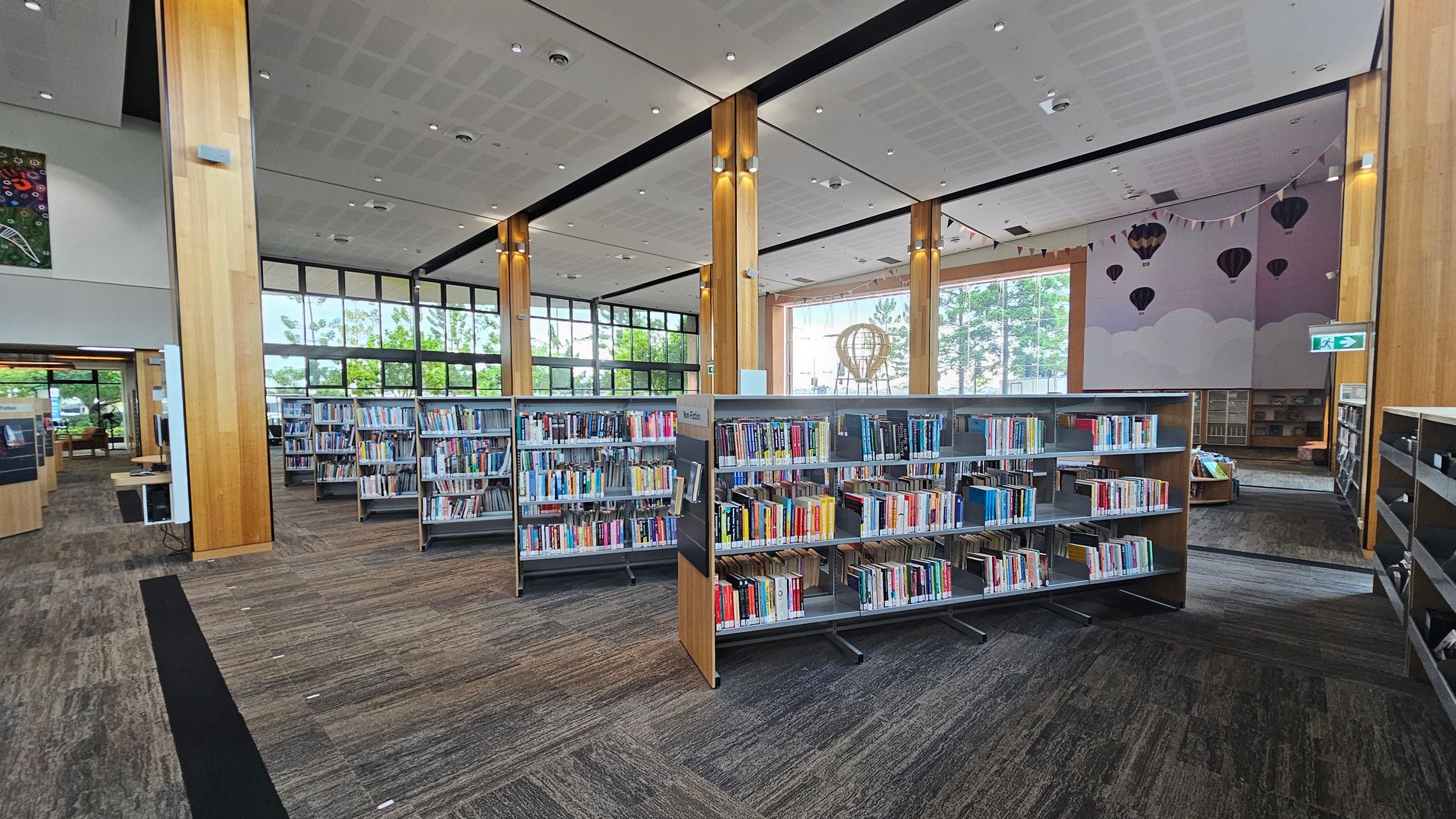 A spacious library interior with tall wooden columns and large windows in the background, allowing natural light to illuminate the space. In the centre, there are several bookshelves filled with books. On the right, a wall features a mural of hot air balloons and clouds, with a small reading area visible. The floor is carpeted in a mix of grey and brown tones, and the ceiling is white with recessed lighting. An exit sign is visible on the right.