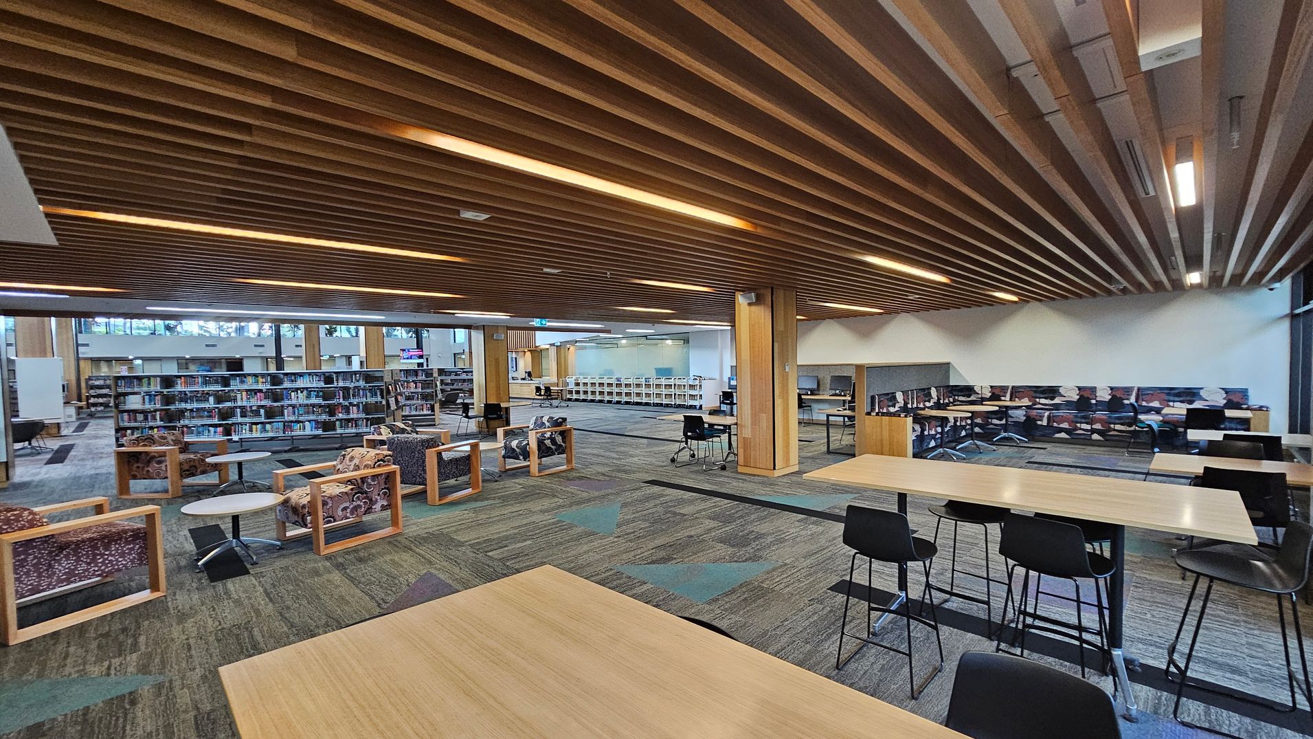 A spacious library interior with a wooden slatted ceiling and patterned carpet flooring. On the left, there are bookshelves filled with books and several cushioned chairs with wooden frames around small round tables. In the centre, a large open area with more seating and tables. On the right, high tables with black chairs and a row of study cubicles along the wall. The background features large windows allowing natural light to enter.