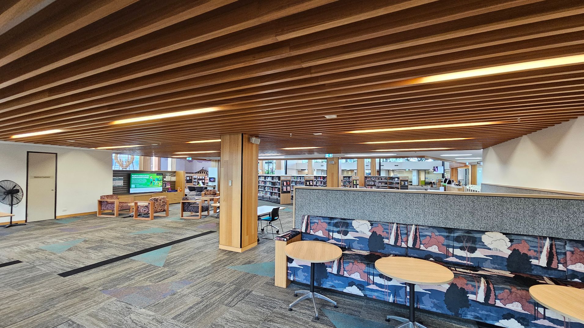 A library interior with a wooden slatted ceiling and patterned carpet flooring. On the left, there is a door and a fan, with a seating area featuring cushioned chairs and a digital display. In the centre, wooden columns support the ceiling, and bookshelves are visible in the background. On the right, a colourful upholstered bench with a nature motif is accompanied by round tables. The space is open and well-lit, with additional seating and bookshelves in the distance.