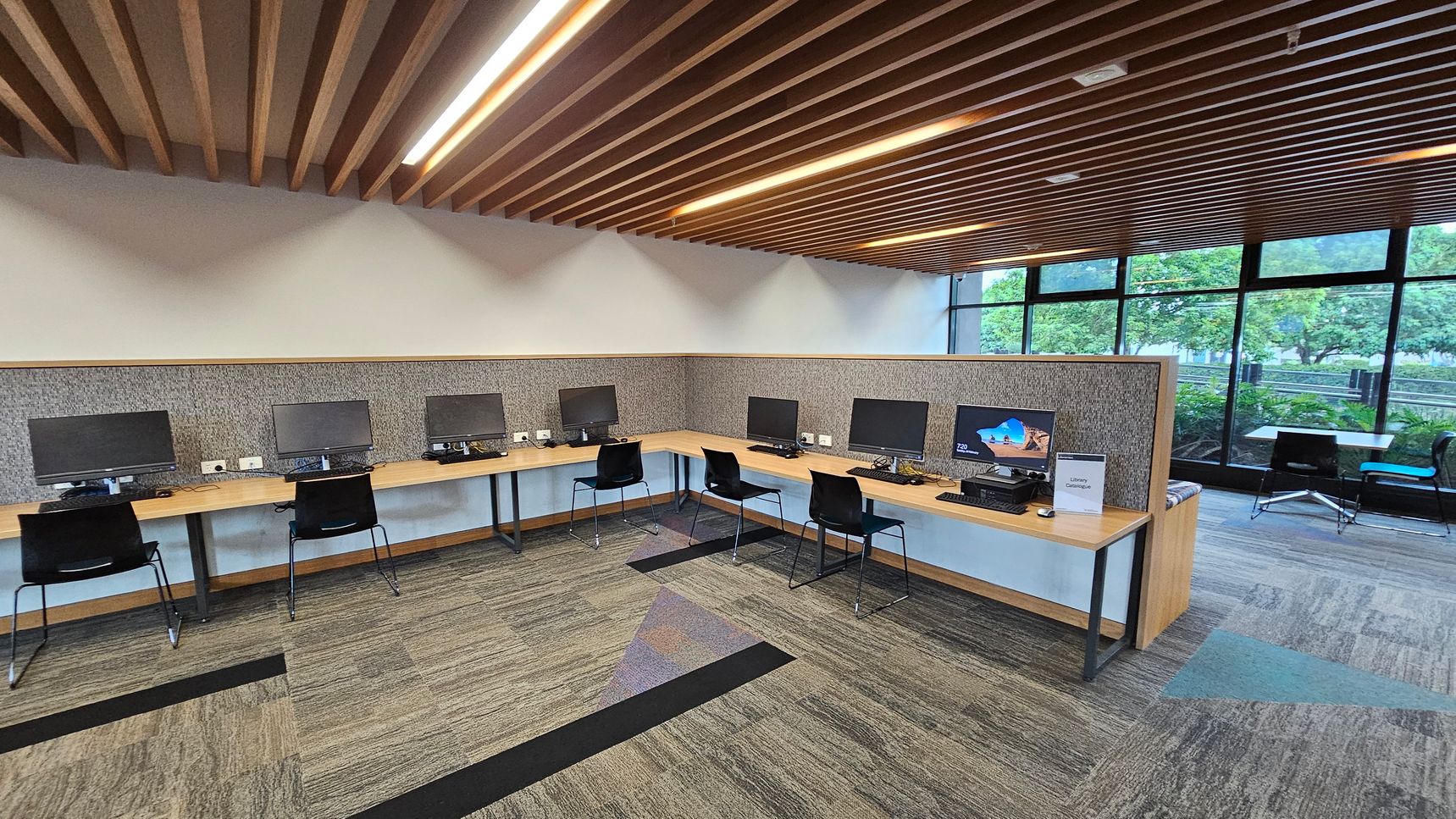 A modern computer lab with a row of desks along the left and back walls, each equipped with a computer monitor and a black chair. The floor is carpeted with a pattern of grey and green tones. The ceiling features wooden slats with integrated lighting. On the right, large windows provide a view of greenery outside. In the foreground, a sign on one of the desks reads "Library Catalogue."