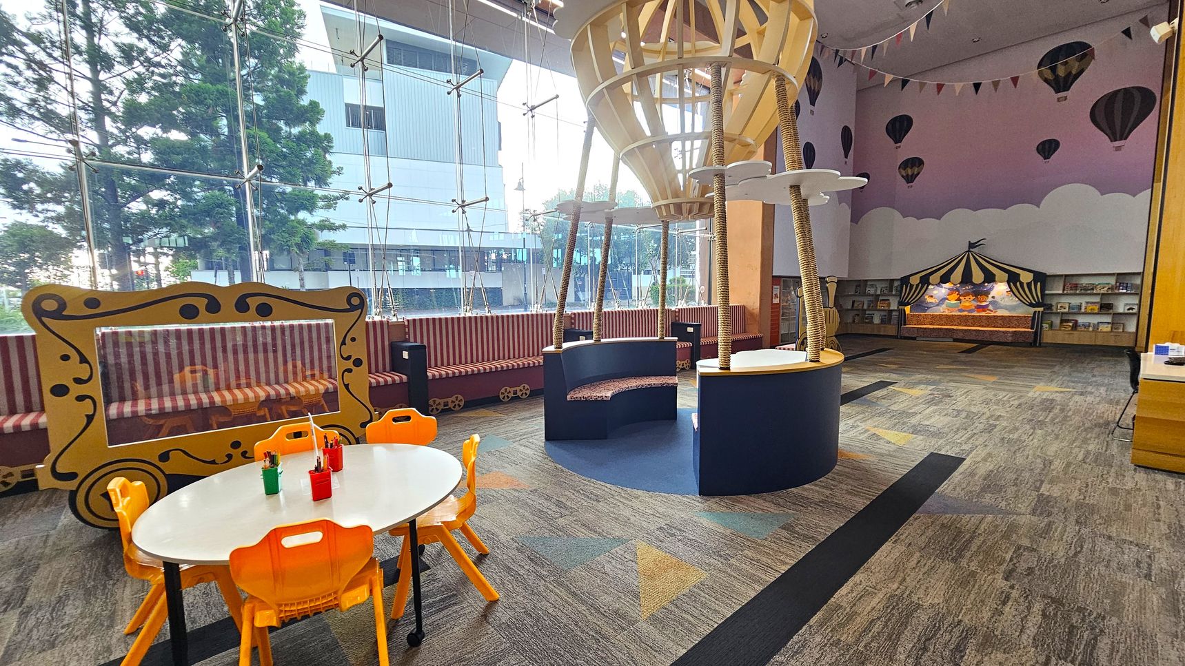 A children's play area with a whimsical design. On the left, there's a white oval table with orange chairs and containers holding crayons. Behind it, a yellow frame resembling a carriage with a striped bench. In the centre, a large hot air balloon structure with seating. The right side features a wall with painted clouds and hot air balloons, a bookshelf, and a circus tent display. The floor is carpeted with geometric patterns, and the ceiling is decorated with bunting. Large windows provide natural light.
