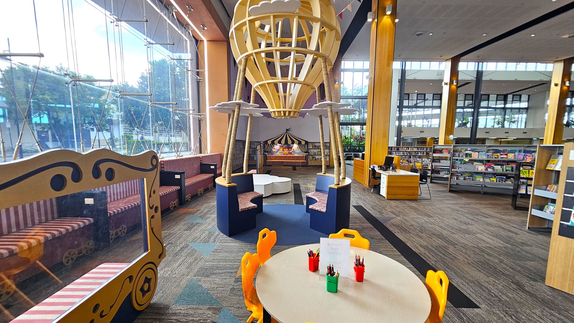 A children's library area with a whimsical design. On the left, there are striped benches with a decorative frame resembling a train carriage. In the centre, a large wooden structure shaped like a hot air balloon with seating underneath. In the foreground, a round table with orange chairs and pots of coloured pencils. On the right, bookshelves filled with books and a wooden desk. The floor is carpeted with a geometric pattern, and the ceiling is high with large windows letting in natural light.