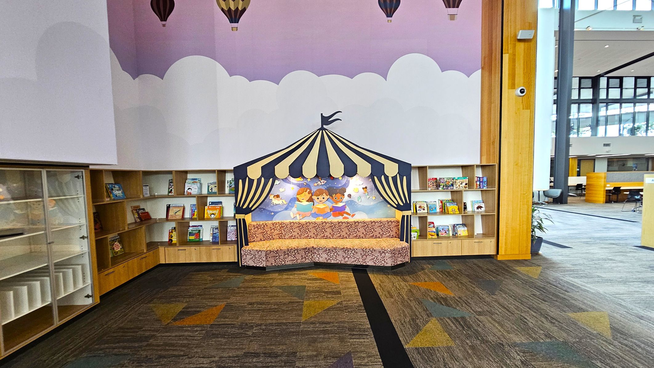 A children's reading area with a colourful, tent-like structure in the centre, featuring an illustration of three children reading. Surrounding the tent are wooden bookshelves filled with children's books. The floor has a patterned carpet with geometric shapes in various colours. The walls are decorated with a mural of clouds and hot air balloons. To the right, there is a large window allowing natural light into the space.