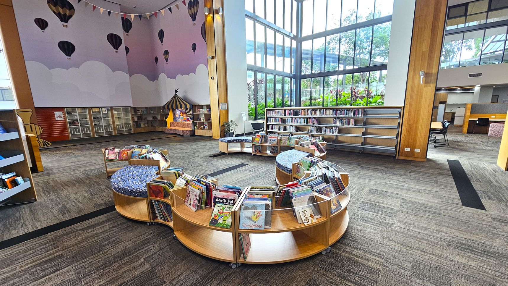 A spacious library area with large floor-to-ceiling windows on the right, allowing natural light to illuminate the space. In the centre, there are circular wooden bookshelves filled with children's books, featuring cushioned seating on top. The left wall is decorated with a mural of hot air balloons and clouds, and there are additional bookshelves and a small reading nook resembling a circus tent. The floor is carpeted in a mix of grey and brown tones.