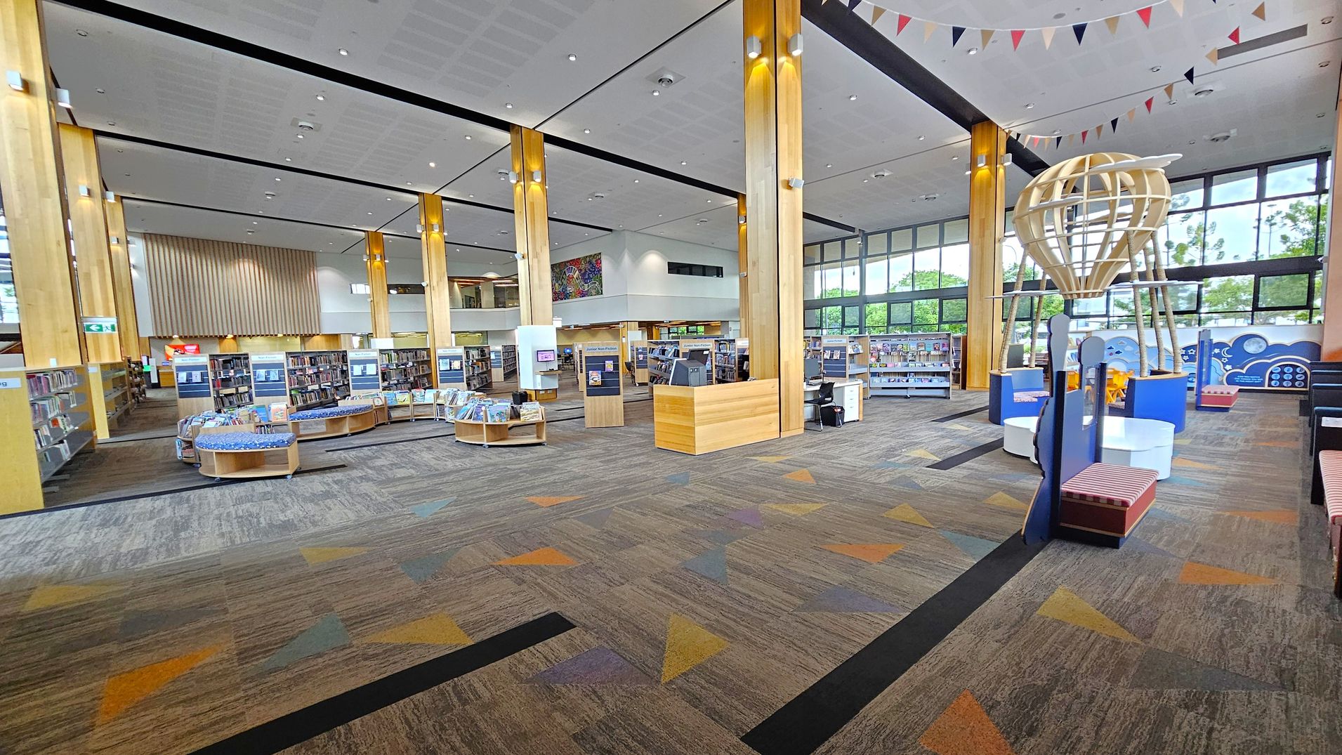 A spacious library interior with a high ceiling and large windows in the background. The floor is carpeted with a geometric pattern in brown, orange, and grey. Wooden bookshelves are arranged in the centre, filled with books. On the right, there's a wooden structure resembling a hot air balloon. Colourful bunting hangs from the ceiling. The left side features more bookshelves and a small desk area. The space is well-lit with natural light and ceiling lights.
