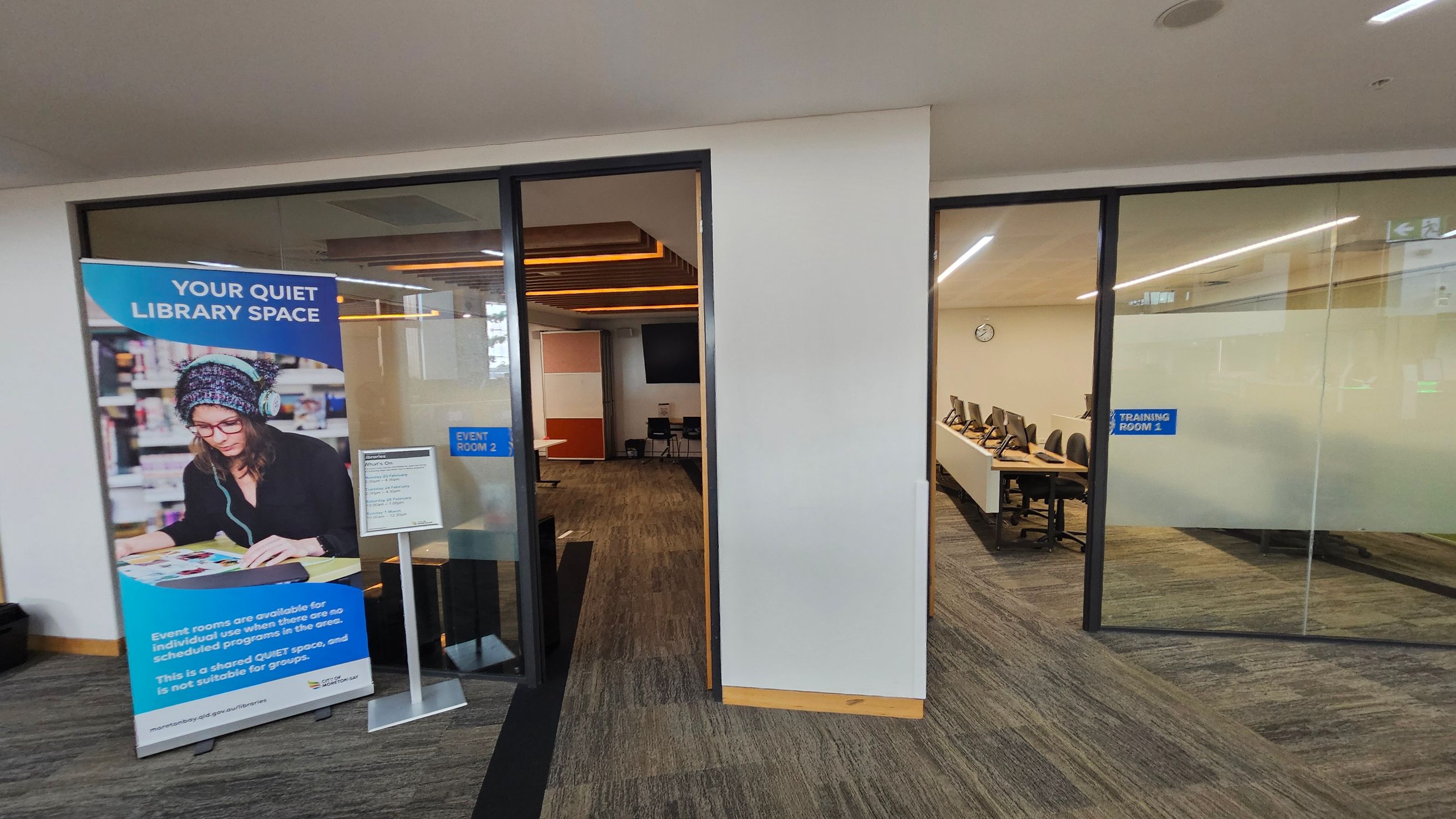 On the left, a large poster displays a person wearing headphones and reading, with text about a quiet library space. Below the poster, a sign lists events. Behind, a glass door leads to "Event Room 2," showing a room with chairs and a screen. On the right, another glass door opens to "Training Room 1," featuring a row of computers on desks. The floor is carpeted in a mix of brown and green, and the ceiling has recessed lighting.