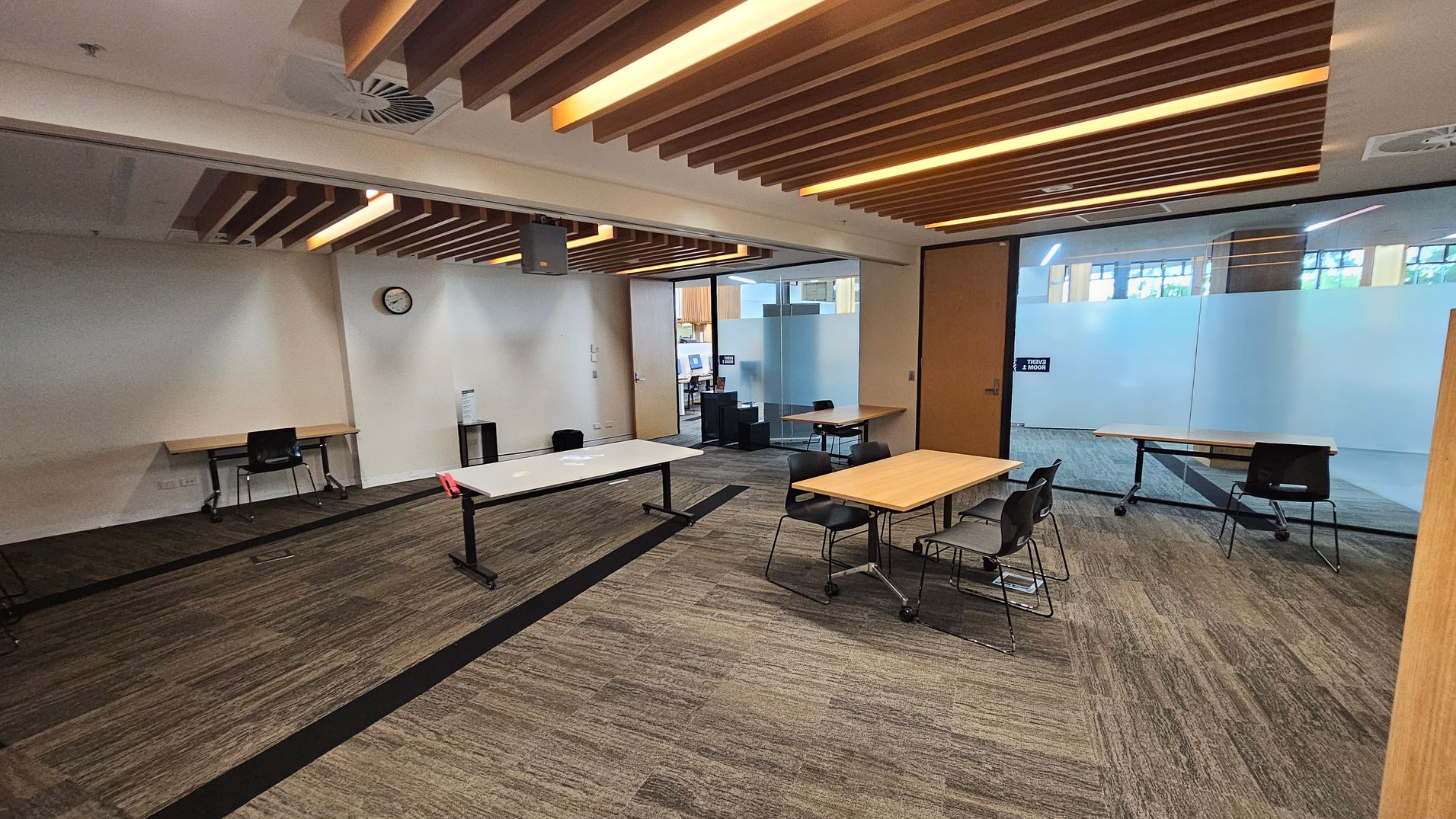 A modern office space with a brown carpeted floor and a ceiling featuring wooden slats with integrated lighting. On the left, a wall-mounted clock is above a desk with a chair. In the centre, there are several tables and chairs, some on wheels. The right side has a large glass partition with frosted sections, and a door with a sign. In the background, there are more desks and chairs visible through the glass.