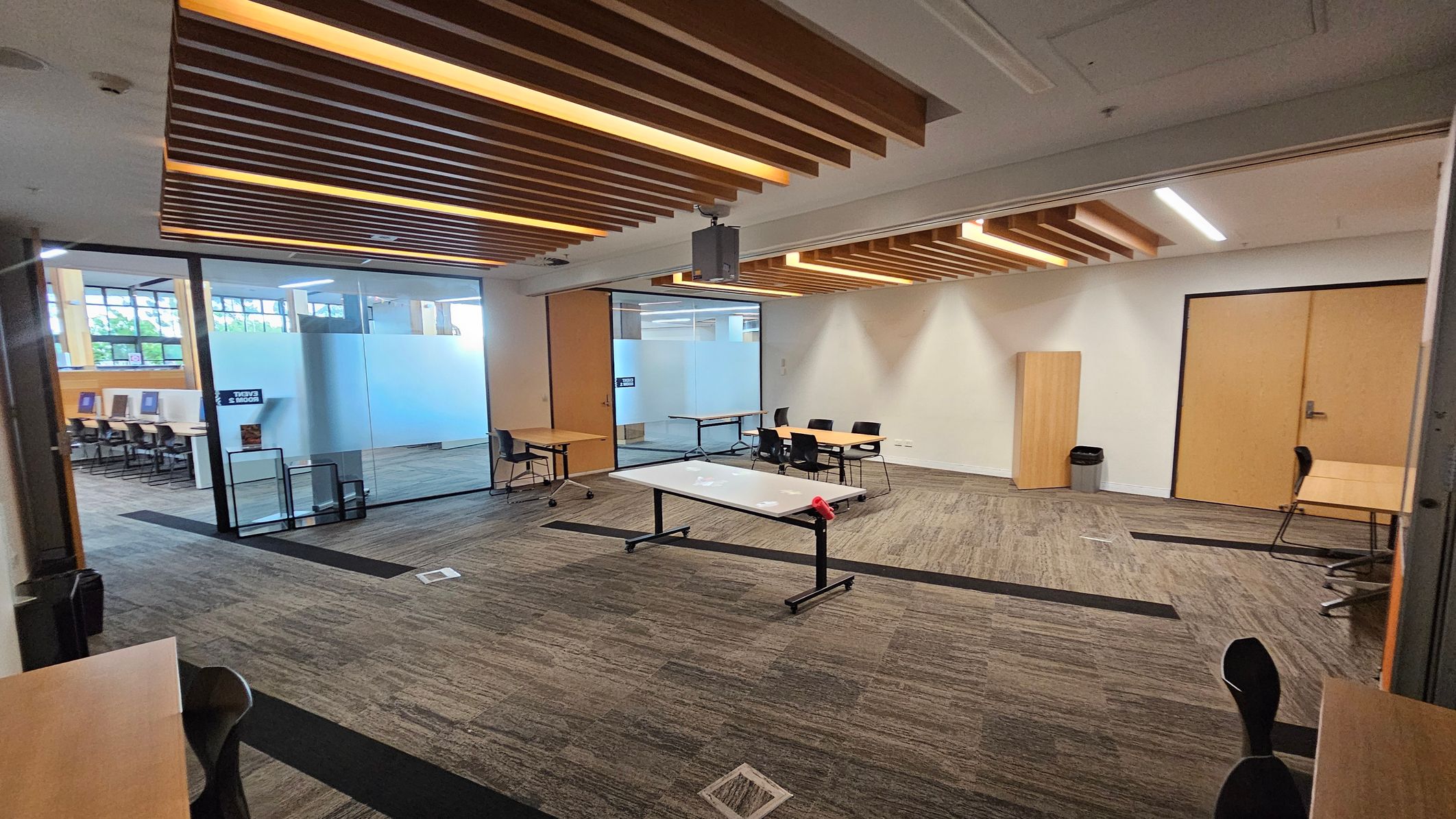 A modern office space with a grey carpeted floor and a ceiling featuring wooden slats with integrated lighting. In the centre, there's a table with a red object on it. To the right, there are wooden doors and a small cabinet with a bin beside it. On the left, a glass partition separates this area from another room with desks and chairs. The background shows large windows allowing natural light in.