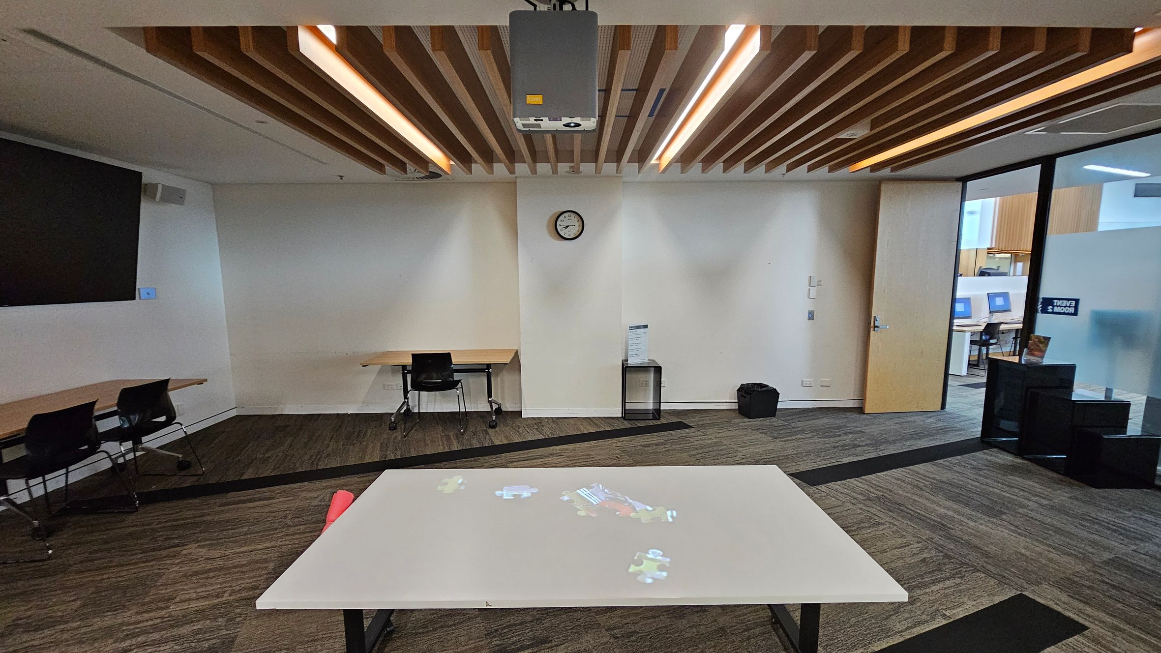 A room with a modern design featuring a wooden slatted ceiling with integrated lighting. In the centre, a white table with a projection on its surface. The back wall has a clock and a small desk with a chair. To the right, a wooden door and a glass partition with a view into another room with computers. The left side has a large wall-mounted screen and a table with chairs. The floor is carpeted with a mix of brown and green patterns.