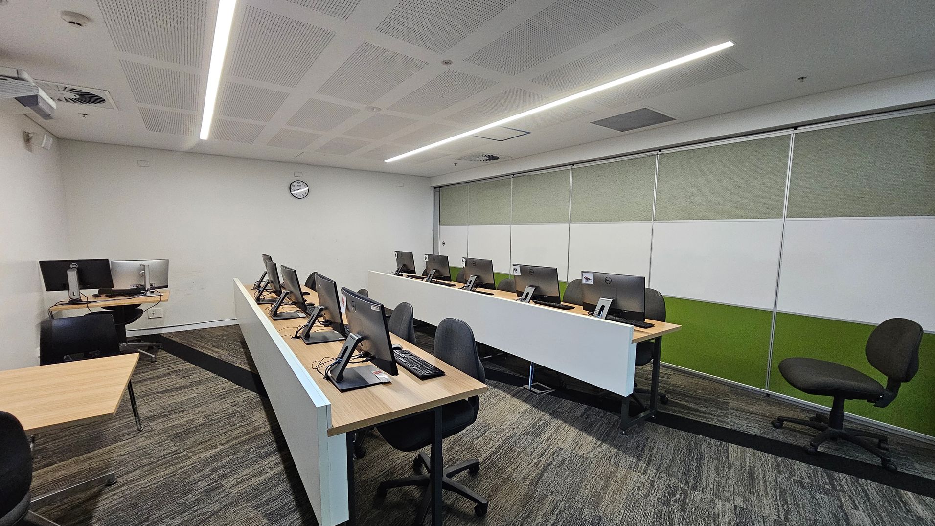 A modern computer lab with two rows of desks, each equipped with computers and black chairs. The left side has a single desk with a computer and chair. The right side features a wall with green and white panels. The floor is carpeted in a mix of green and grey patterns. The ceiling is white with recessed lighting and ventilation panels. A clock is mounted on the back wall.