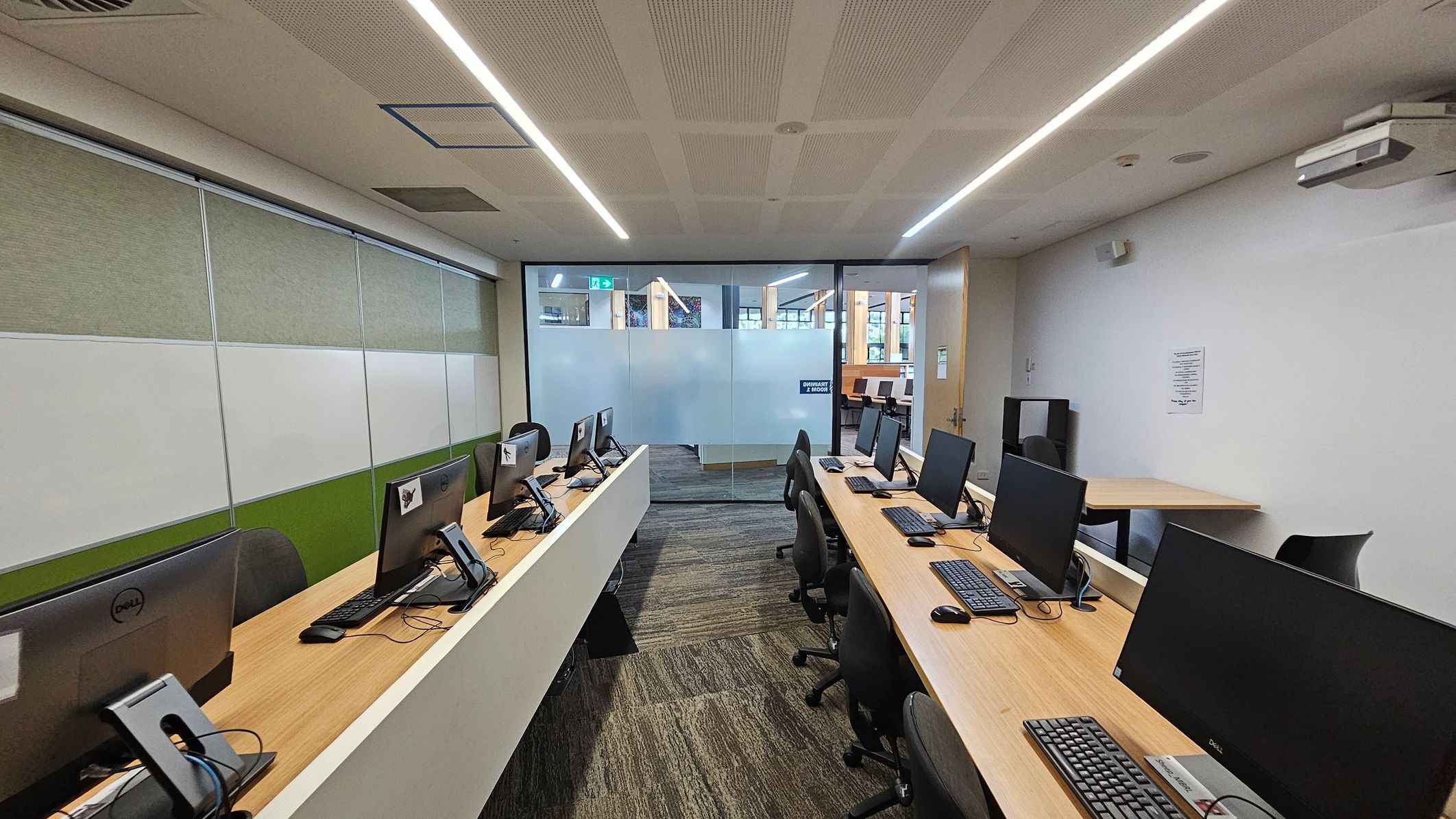A computer lab with two rows of desks, each equipped with desktop computers and chairs. The left wall has green and white panels, while the right wall is plain with a notice. The floor is carpeted in a green and brown pattern. At the back, there is a glass partition with frosted sections, leading to another room. The ceiling features recessed lighting and ventilation panels.