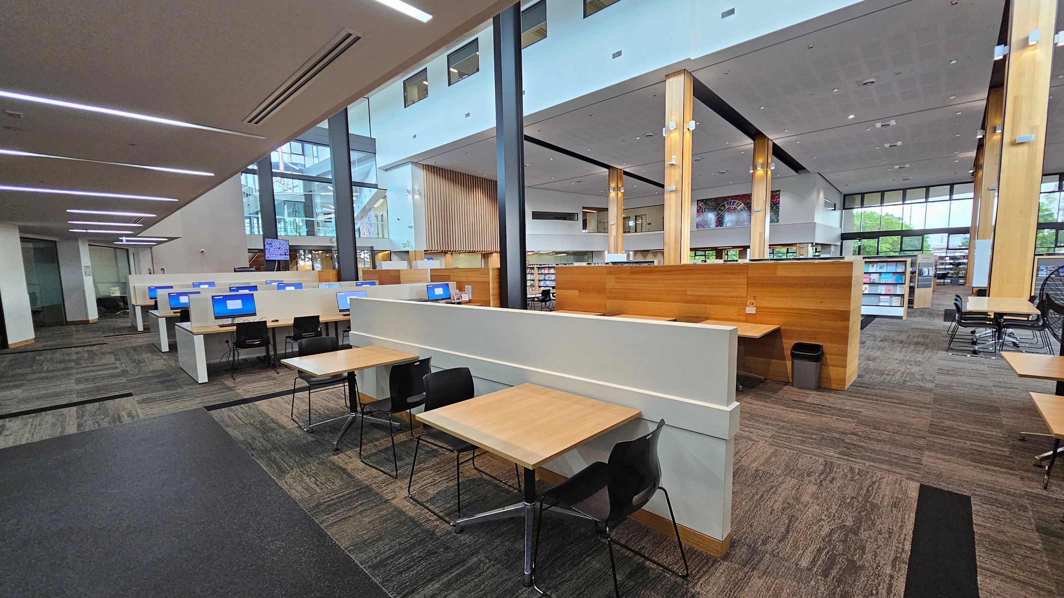 A modern library interior with a spacious layout. On the left, there are computer workstations with blue screens and black chairs. In the centre, wooden tables with black chairs are arranged for study. The right side features bookshelves and additional seating. The floor is carpeted in a mix of grey and green tones. The ceiling is high with recessed lighting, and large windows provide natural light. Wooden pillars and partitions add warmth to the space.