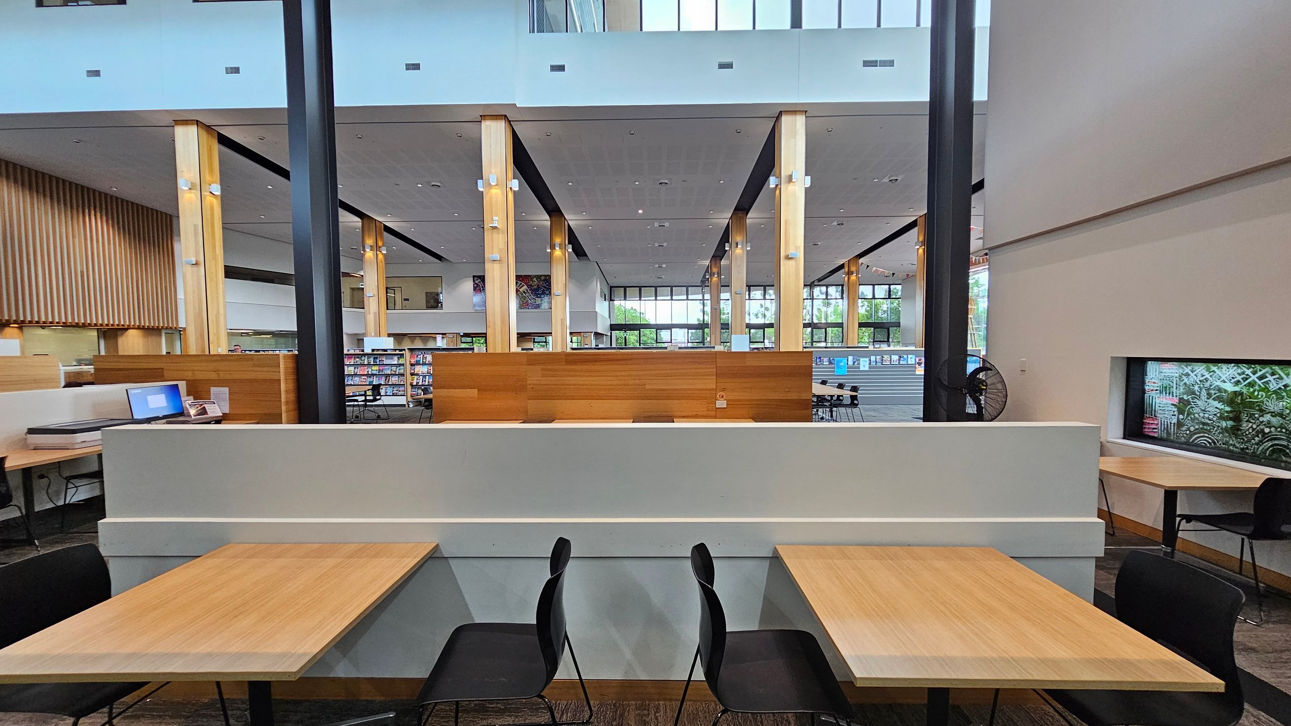 In the foreground, there are two wooden tables with black chairs. A grey partition separates the tables from the rest of the space. In the background, the area is open with wooden columns and a high ceiling. There are bookshelves and a large window allowing natural light. On the left, a computer is on a desk, and on the right, a window with a view of greenery. The floor is carpeted, and the ceiling is dotted with lights.