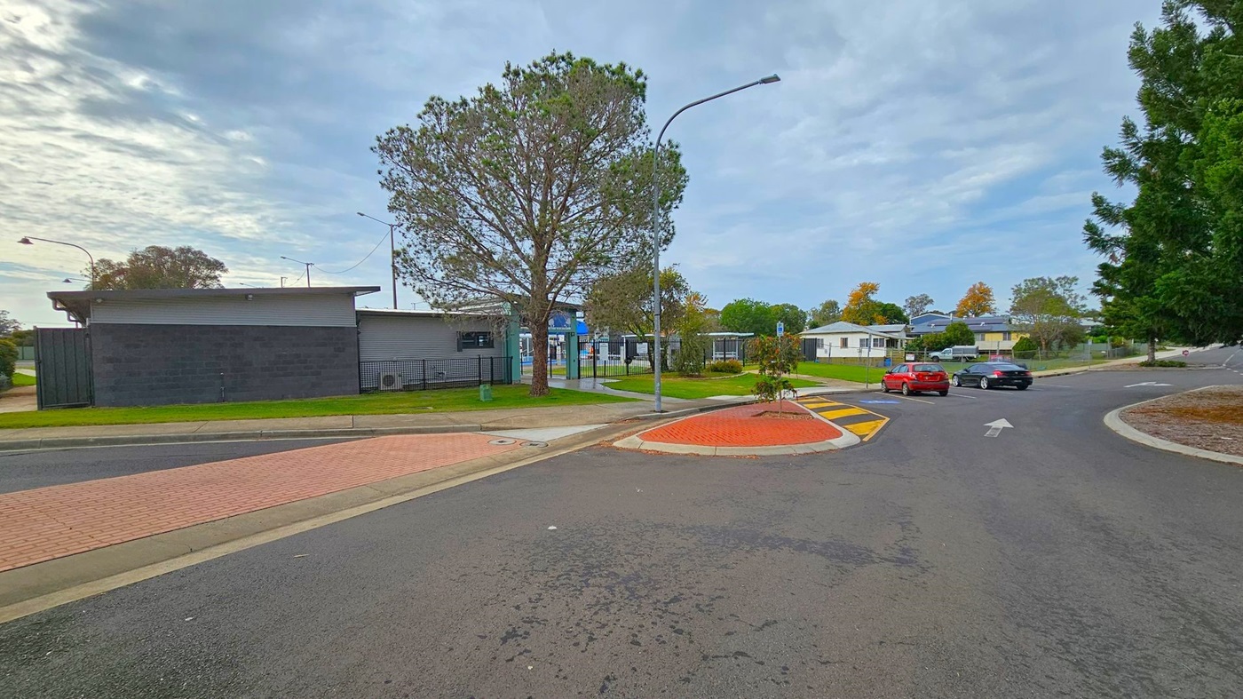 A street scene with a black building on the left, featuring a flat roof and surrounded by a grassy area. A large tree stands nearby, next to a metal fence. In the centre, a road curves to the right, with a red car and a black car driving away. On the right, there are more trees and a few residential buildings in the background. The road has a yellow and black speed bump and a red brick pedestrian crossing. Street lamps are visible along the road. A street scene with a black building on the left, featuring a flat roof and surrounded by a grassy area. A large tree stands nearby, next to a metal fence. In the centre, a road curves to the right, with a red car and a black car driving away. On the right, there are more trees and a few residential buildings in the background. The road has a yellow and black speed bump and a red brick pedestrian crossing. Street lamps are visible along the road.