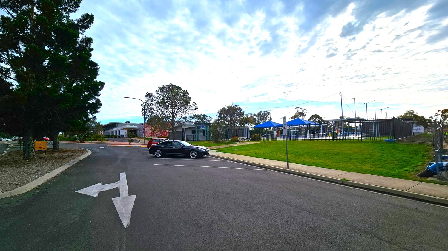 A road with a white directional arrow painted on it. On the left, there are tall trees and a small building in the distance. A black car is parked near the centre, with a grassy area and a fence to the right. Behind the fence, there are blue canopies and more trees. The sky is overcast with scattered clouds. A road with a white directional arrow painted on it. On the left, there are tall trees and a small building in the distance. A black car is parked near the centre, with a grassy area and a fence to the right. Behind the fence, there are blue canopies and more trees. The sky is overcast with scattered clouds.