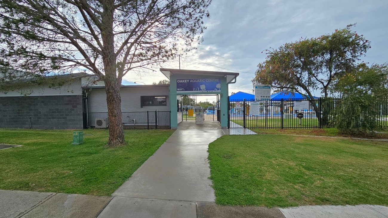 A pathway leads to the entrance of the Oakey Aquatic Centre, flanked by a large tree on the left and a smaller tree on the right. The building on the left has a grey exterior with a window. A black metal fence surrounds the area, with signs visible near the entrance. Blue shade structures can be seen in the background, indicating a pool area. The ground is covered with grass and concrete paths. A pathway leads to the entrance of the Oakey Aquatic Centre, flanked by a large tree on the left and a smaller tree on the right. The building on the left has a grey exterior with a window. A black metal fence surrounds the area, with signs visible near the entrance. Blue shade structures can be seen in the background, indicating a pool area. The ground is covered with grass and concrete paths.