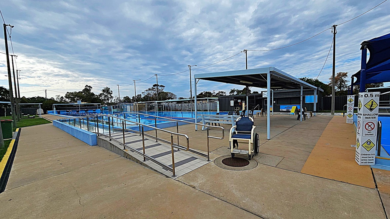 A small outdoor swimming pool is covered by a blue shade structure. The pool has a ramp with handrails for accessibility and a mushroom-shaped water feature. Surrounding the pool is a paved area with grass patches. A wheelchair is parked near the ramp. In the background, there are more pools, benches, and a fence with trees beyond it. The area is well-lit with tall light poles. A small outdoor swimming pool is covered by a blue shade structure. The pool has a ramp with handrails for accessibility and a mushroom-shaped water feature. Surrounding the pool is a paved area with grass patches. A wheelchair is parked near the ramp. In the background, there are more pools, benches, and a fence with trees beyond it. The area is well-lit with tall light poles.