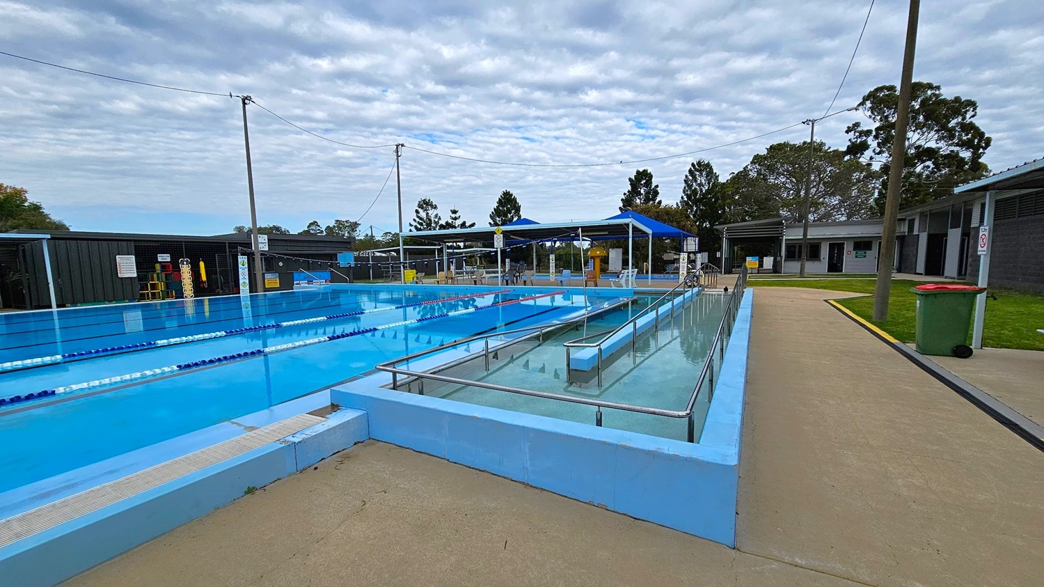 Outdoor swimming pool area with lanes on the left and a ramp leading into the water on the right. The pool is surrounded by a concrete walkway. In the background, there are blue shade structures, tables, and chairs. A building with several doors is visible on the far right. Trees and a grassy area are in the background, and a green bin with a red lid is near the walkway. Overhead wires are strung between poles. Outdoor swimming pool area with lanes on the left and a ramp leading into the water on the right. The pool is surrounded by a concrete walkway. In the background, there are blue shade structures, tables, and chairs. A building with several doors is visible on the far right. Trees and a grassy area are in the background, and a green bin with a red lid is near the walkway. Overhead wires are strung between poles.