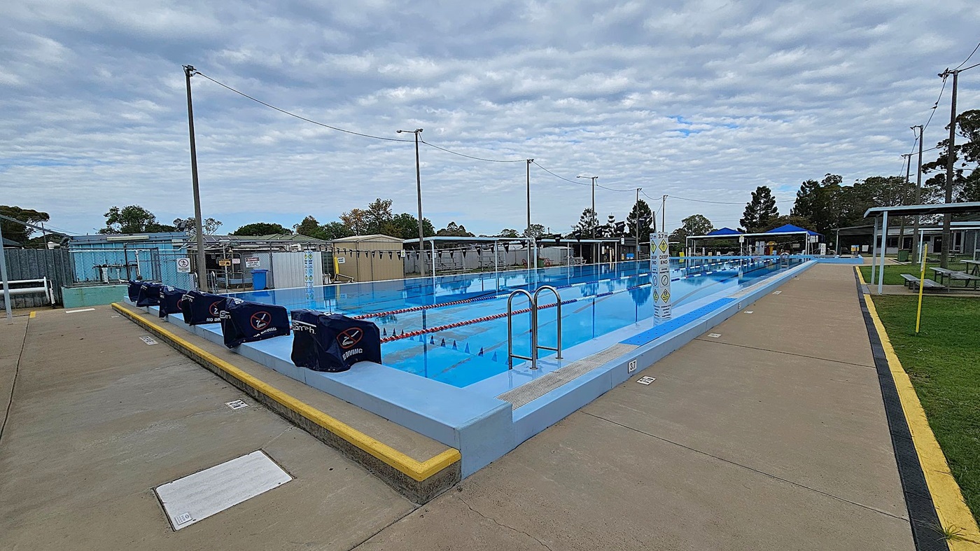 An outdoor swimming pool with multiple lanes is in the centre, surrounded by a concrete walkway. On the left, there are poolside structures and equipment. The right side features a grassy area with a few trees and a fence. In the background, there are small buildings and more trees. Several tall light poles are positioned around the pool area. An outdoor swimming pool with multiple lanes is in the centre, surrounded by a concrete walkway. On the left, there are poolside structures and equipment. The right side features a grassy area with a few trees and a fence. In the background, there are small buildings and more trees. Several tall light poles are positioned around the pool area.