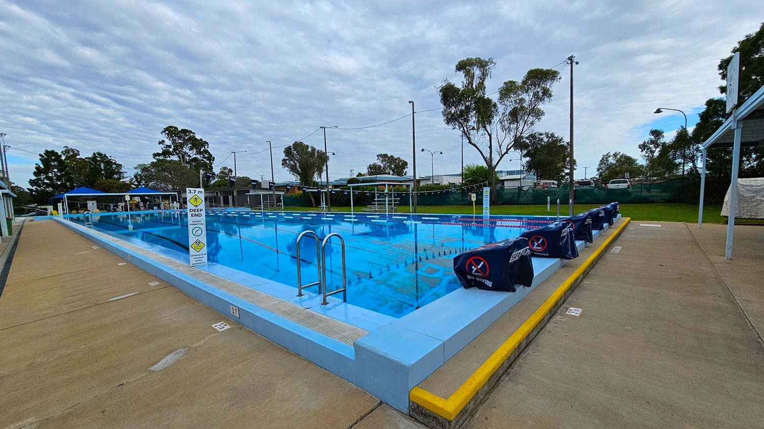 An outdoor swimming pool with clear blue water is in the centre, surrounded by a concrete deck. On the left, there are starting blocks covered with dark blue covers. A metal ladder descends into the pool. The right side features a paved walkway with a grassy area and picnic tables under a shelter. In the background, there are small buildings and trees, with tall light poles lining the pool area. An outdoor swimming pool with clear blue water is in the centre, surrounded by a concrete deck. On the left, there are starting blocks covered with dark blue covers. A metal ladder descends into the pool. The right side features a paved walkway with a grassy area and picnic tables under a shelter. In the background, there are small buildings and trees, with tall light poles lining the pool area.