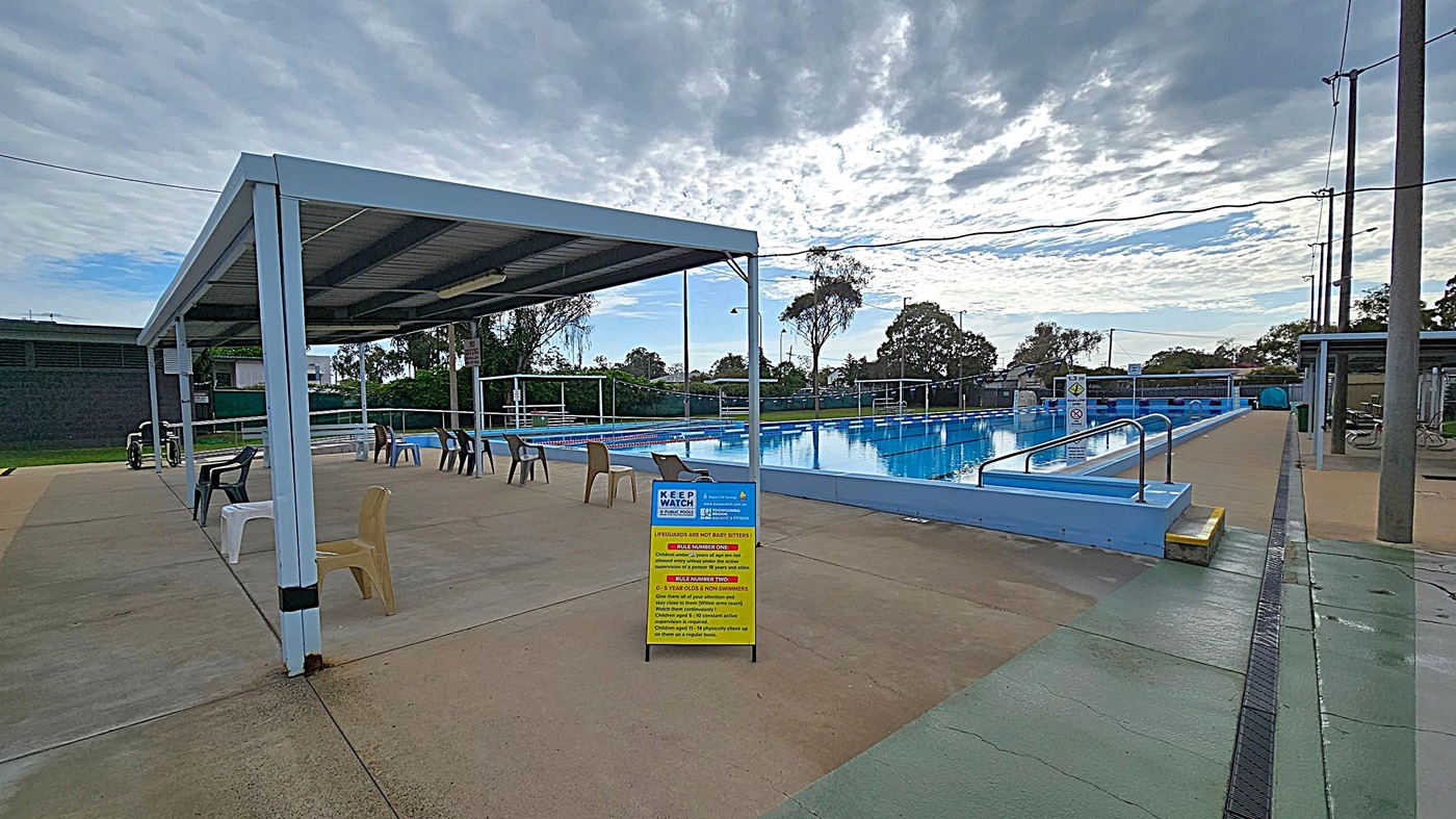 An outdoor swimming pool with clear blue water, surrounded by a concrete deck. On the left, there is a walkway with a sign indicating a depth of 3.7 metres. The right side has a series of covered starting blocks. In the background, there are trees and a few parked cars behind a green fence. The pool is equipped with lane dividers, and there are some blue canopies providing shade over seating areas. An outdoor swimming pool with clear blue water, surrounded by a concrete deck. On the left, there is a walkway with a sign indicating a depth of 3.7 metres. The right side has a series of covered starting blocks. In the background, there are trees and a few parked cars behind a green fence. The pool is equipped with lane dividers, and there are some blue canopies providing shade over seating areas.