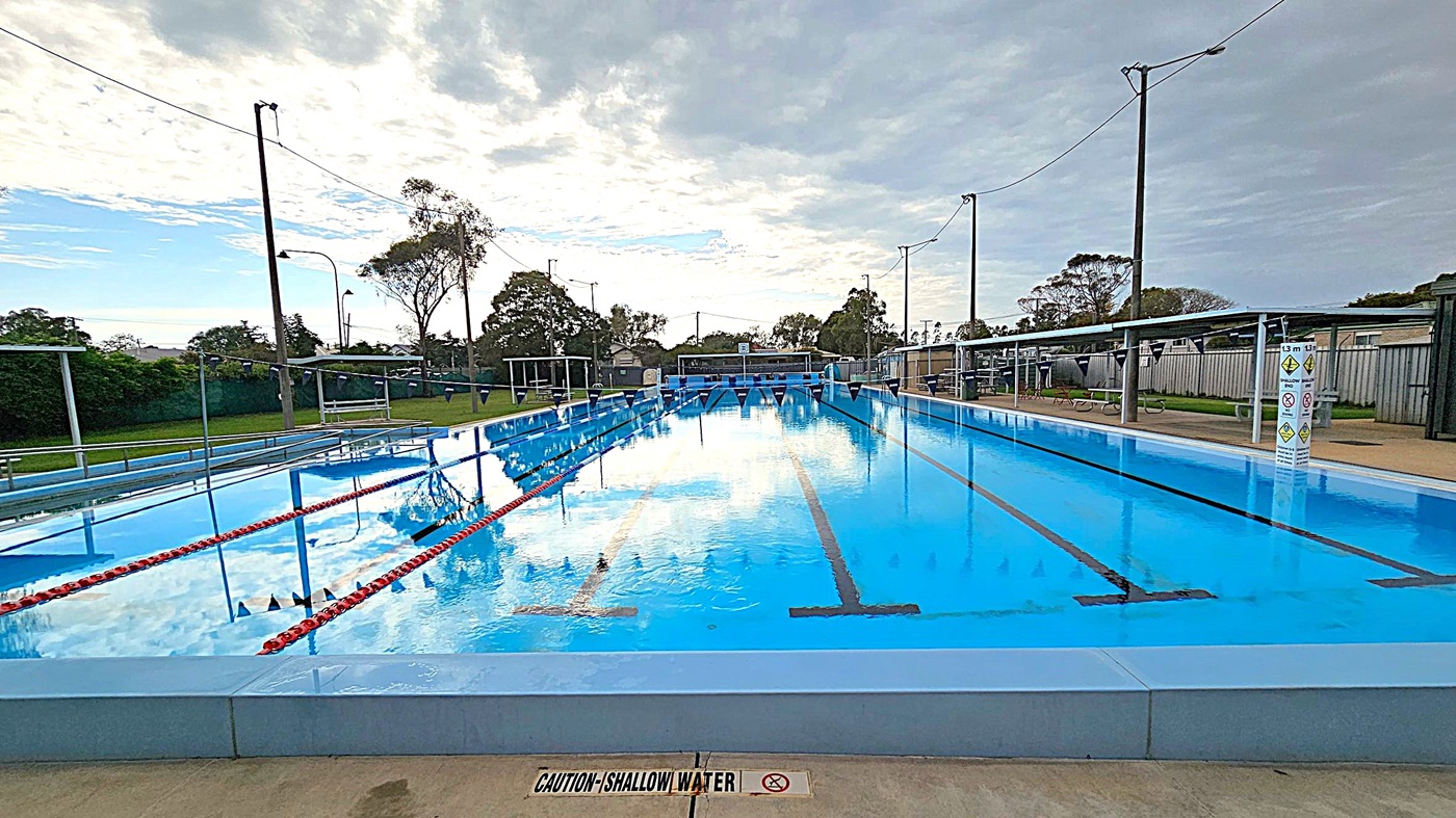 Outdoor swimming pool area with a covered seating section on the left, featuring several plastic chairs. In the centre, a sign with safety instructions is visible. The pool is on the right, with clear water and lane dividers. Trees and some buildings are in the background. The ground is a mix of green and beige concrete, and the pool has metal handrails. The sky is overcast with patches of blue. Outdoor swimming pool area with a covered seating section on the left, featuring several plastic chairs. In the centre, a sign with safety instructions is visible. The pool is on the right, with clear water and lane dividers. Trees and some buildings are in the background. The ground is a mix of green and beige concrete, and the pool has metal handrails. The sky is overcast with patches of blue.